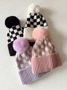 Four stylish beanies featuring pom-poms and checker patterns in various colors: black, pink, and lavender.