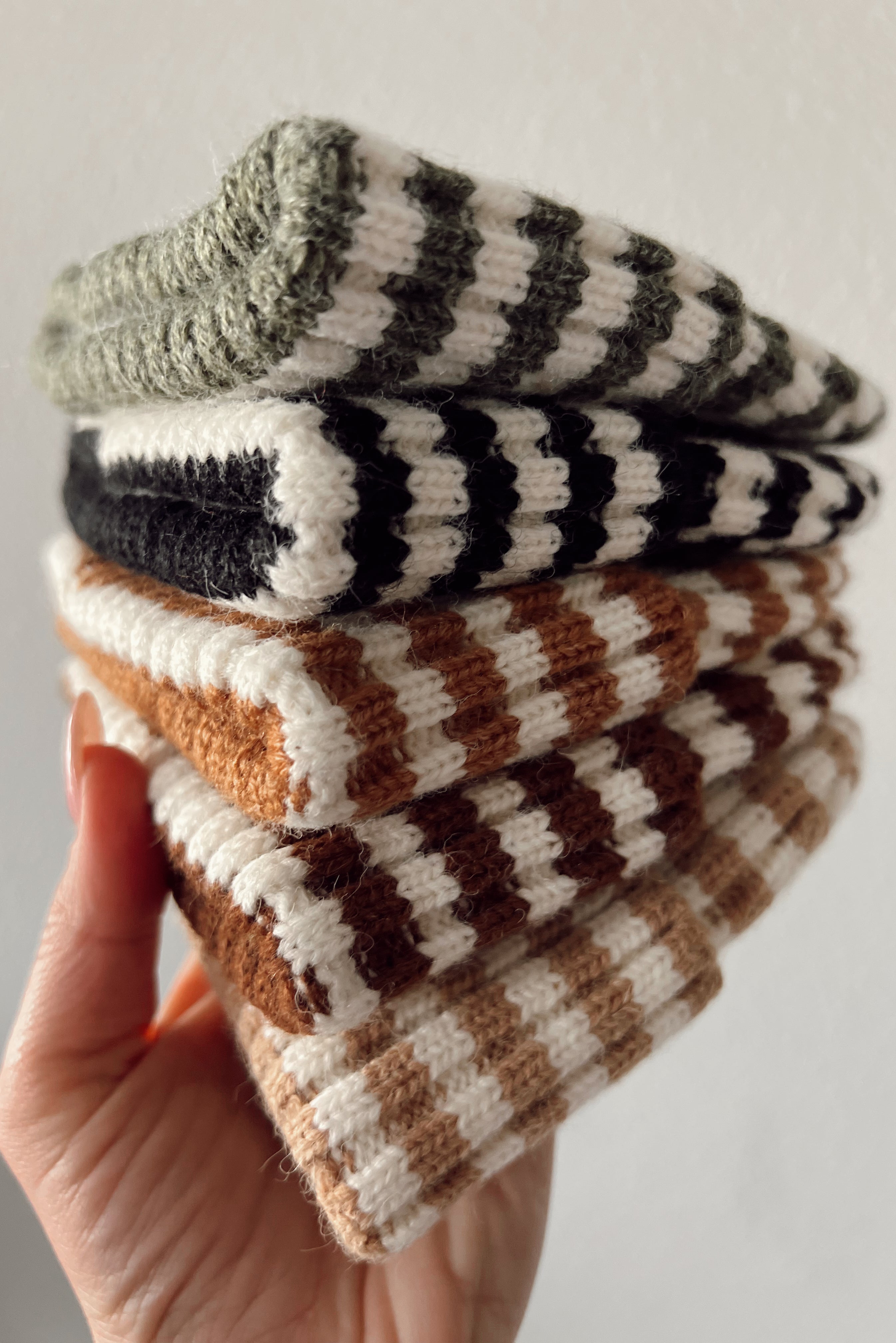 A hand holding a stack of striped knit hats in earthy tones against a neutral background.