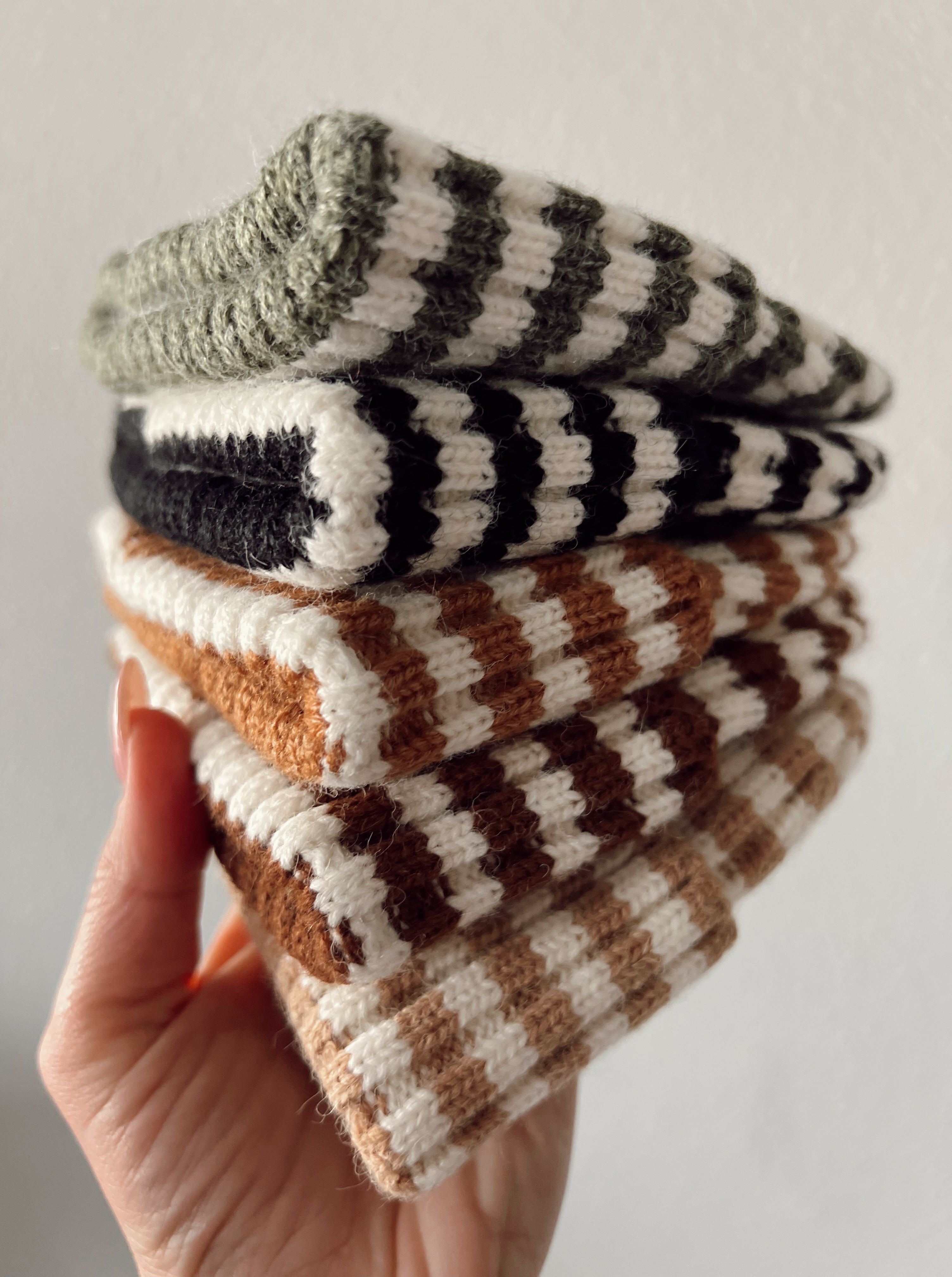 A hand holding a stack of striped knit hats in earthy tones against a neutral background.