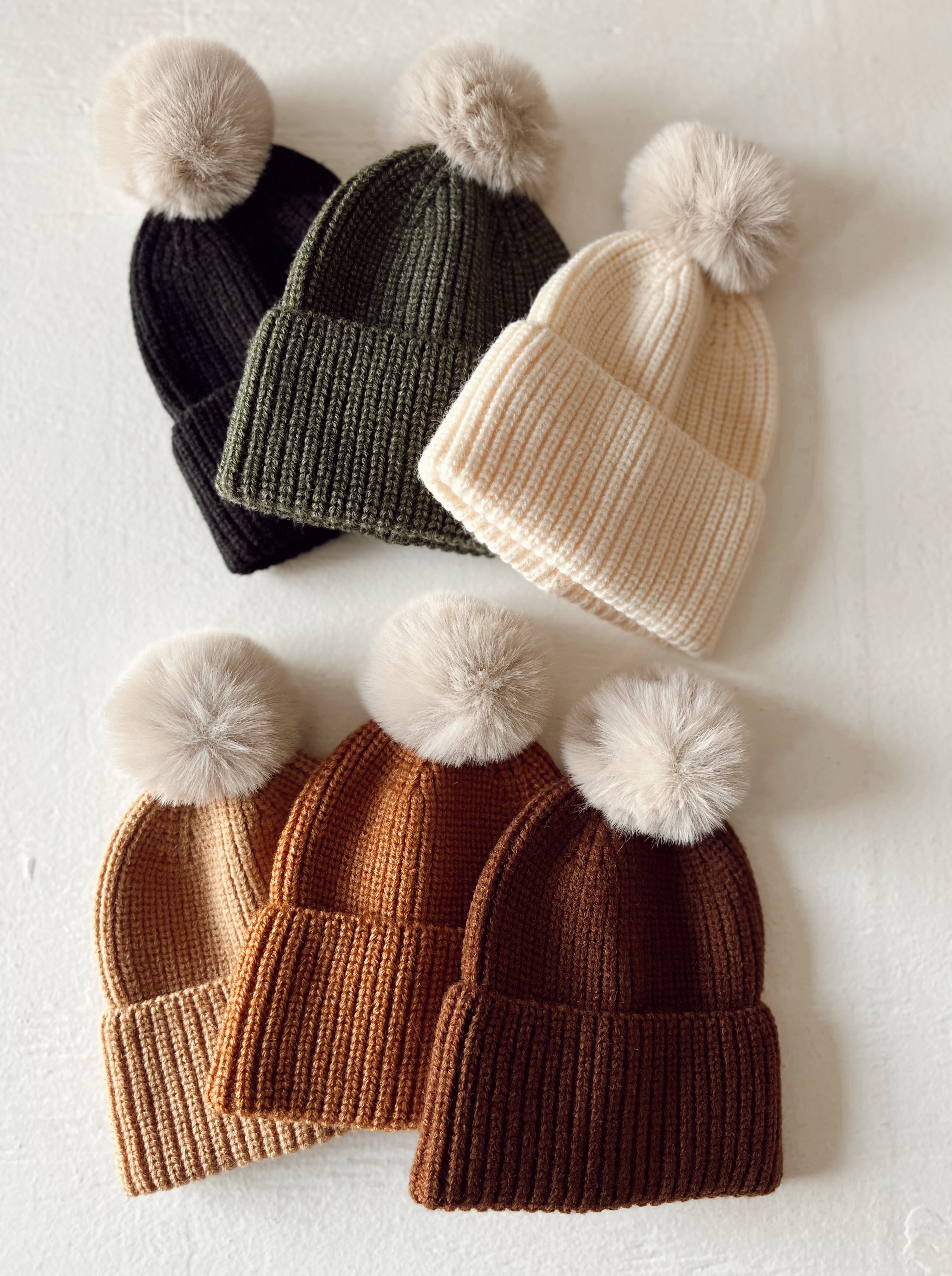 Various knitted beanies with fluffy pom-poms in black, olive, cream, tan, and brown arranged on a light background.