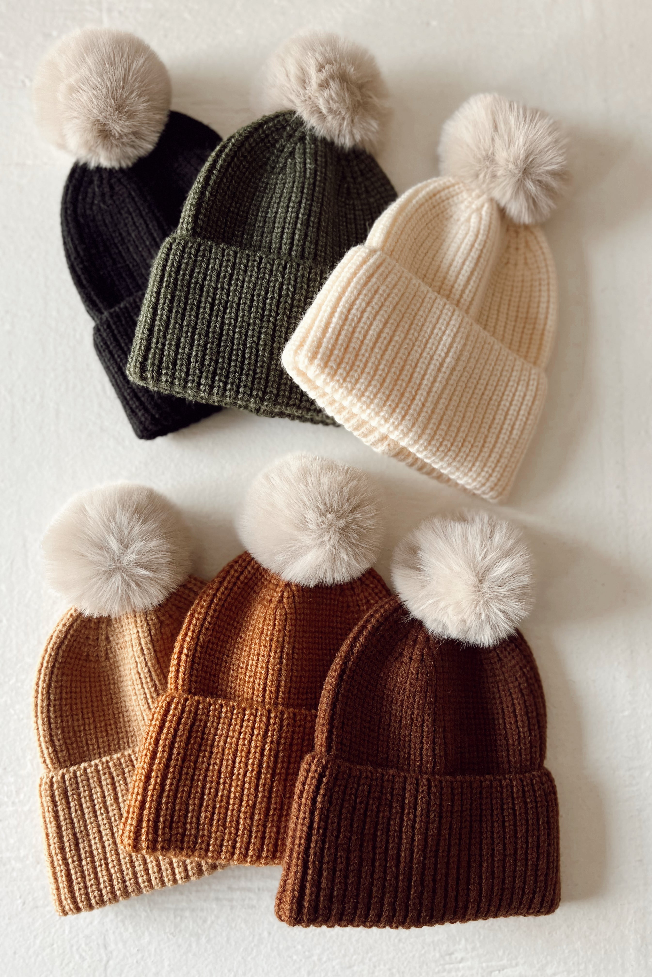 Collection of knit beanie hats in various colors with fluffy pom-poms on top, arranged on a light background.