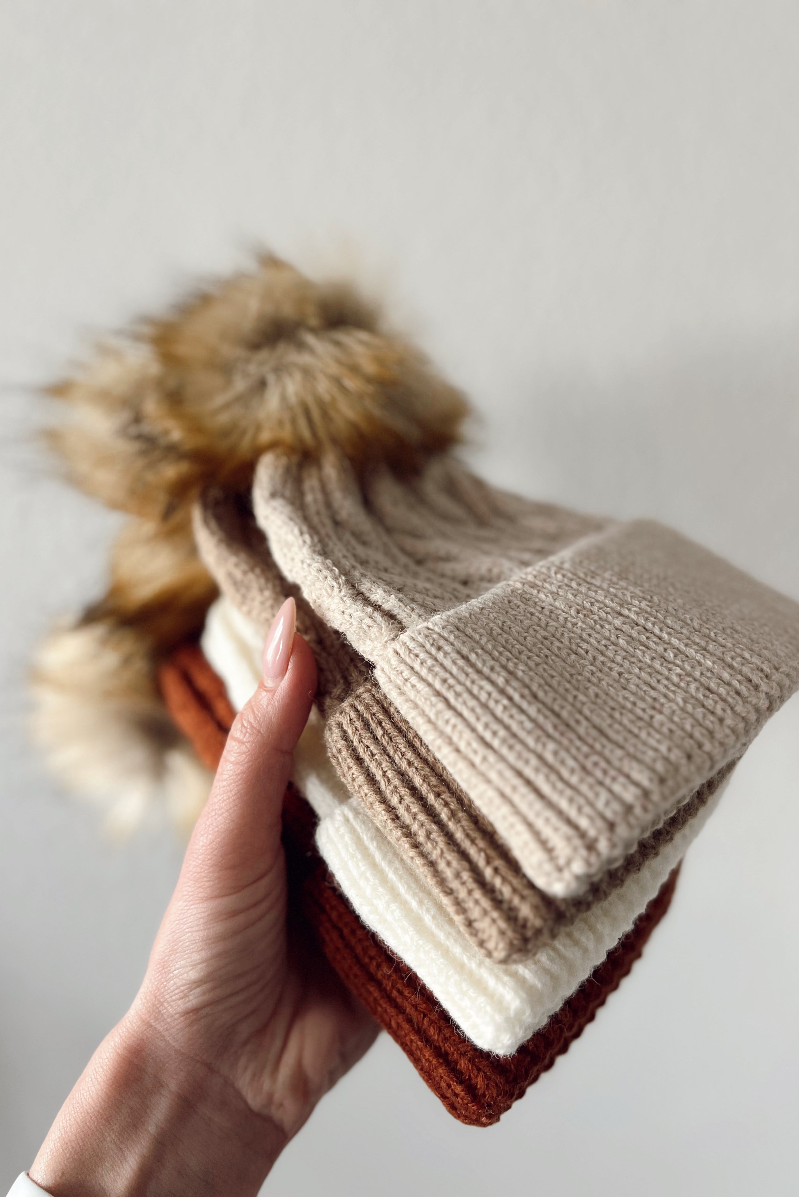 A hand holding four knitted winter hats in cream, beige, brown, and rust, featuring fur pom-poms.