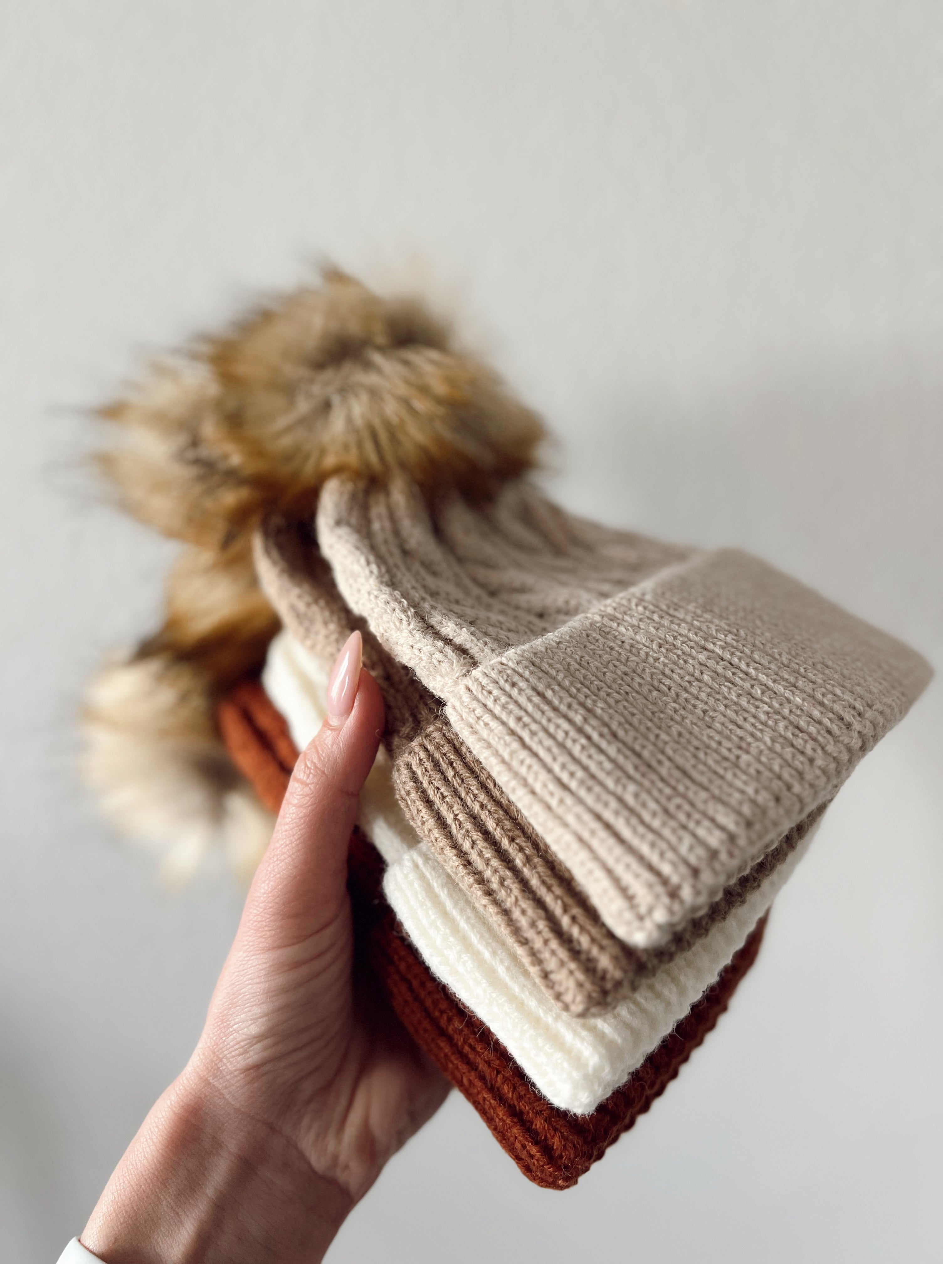 A hand holding four knitted winter hats in cream, beige, brown, and rust, featuring fur pom-poms.