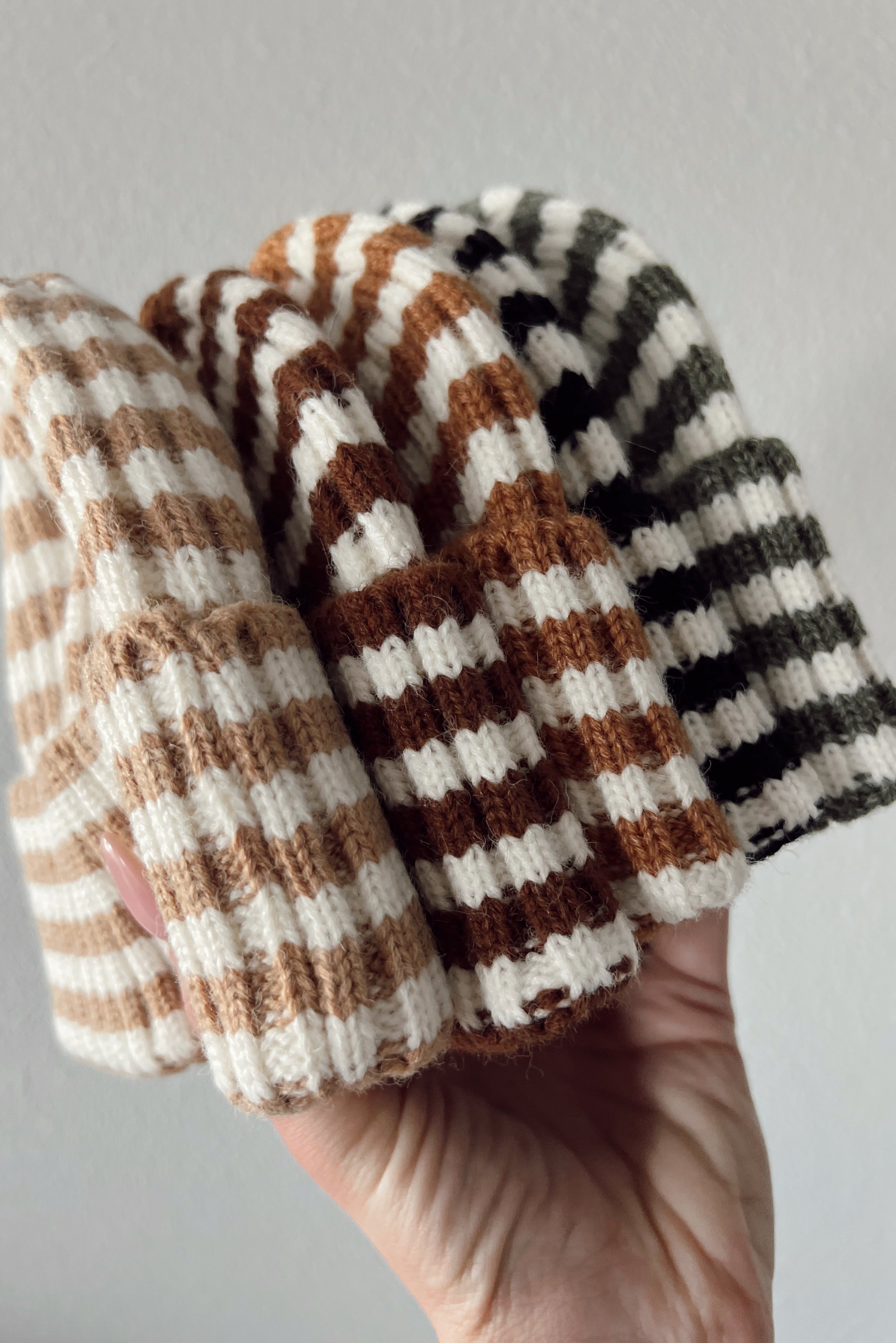 A hand holding four striped beanies in beige, brown, and green colors against a light background.