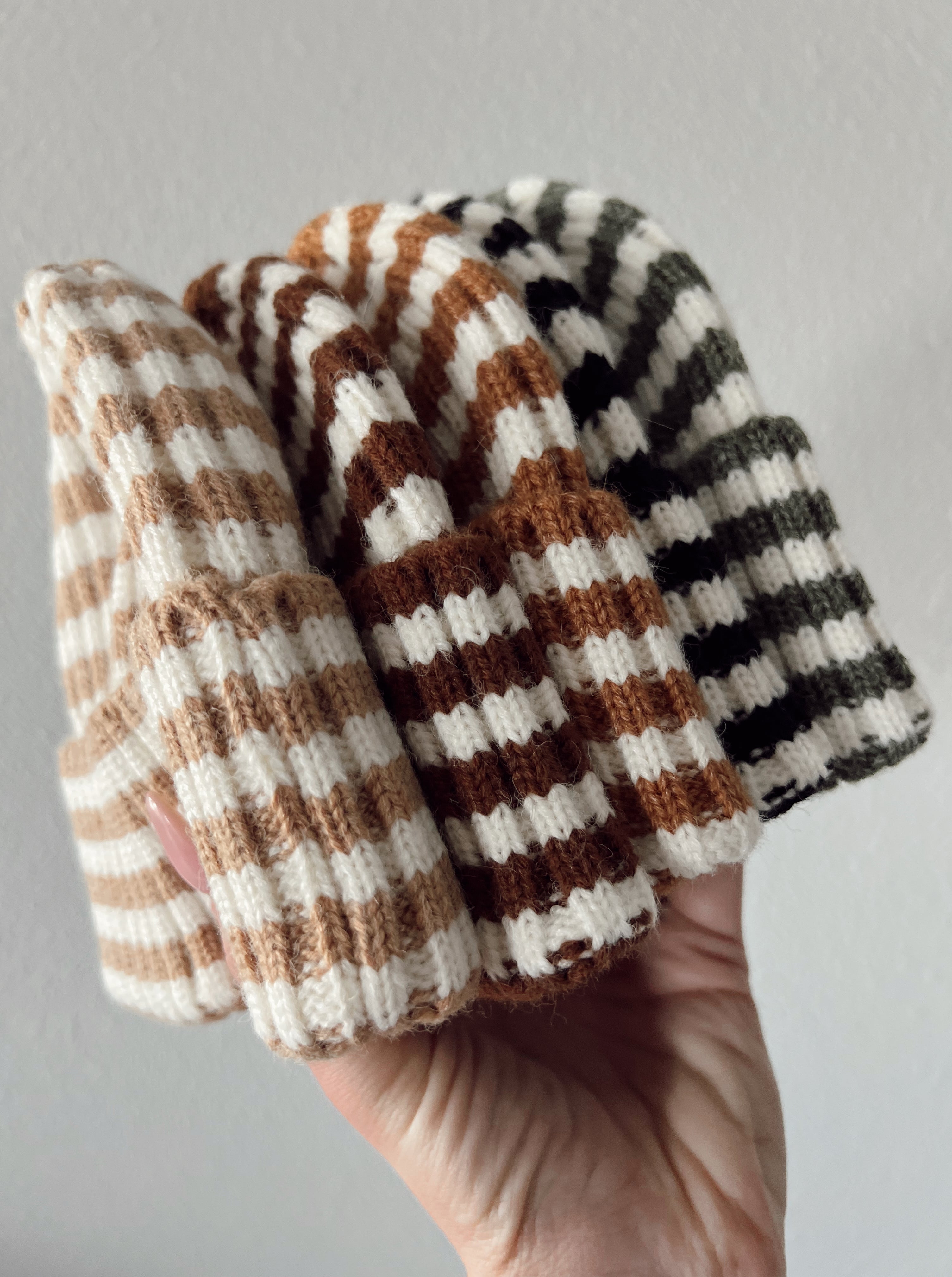 A hand holding four striped beanies in beige, brown, and green colors against a light background.