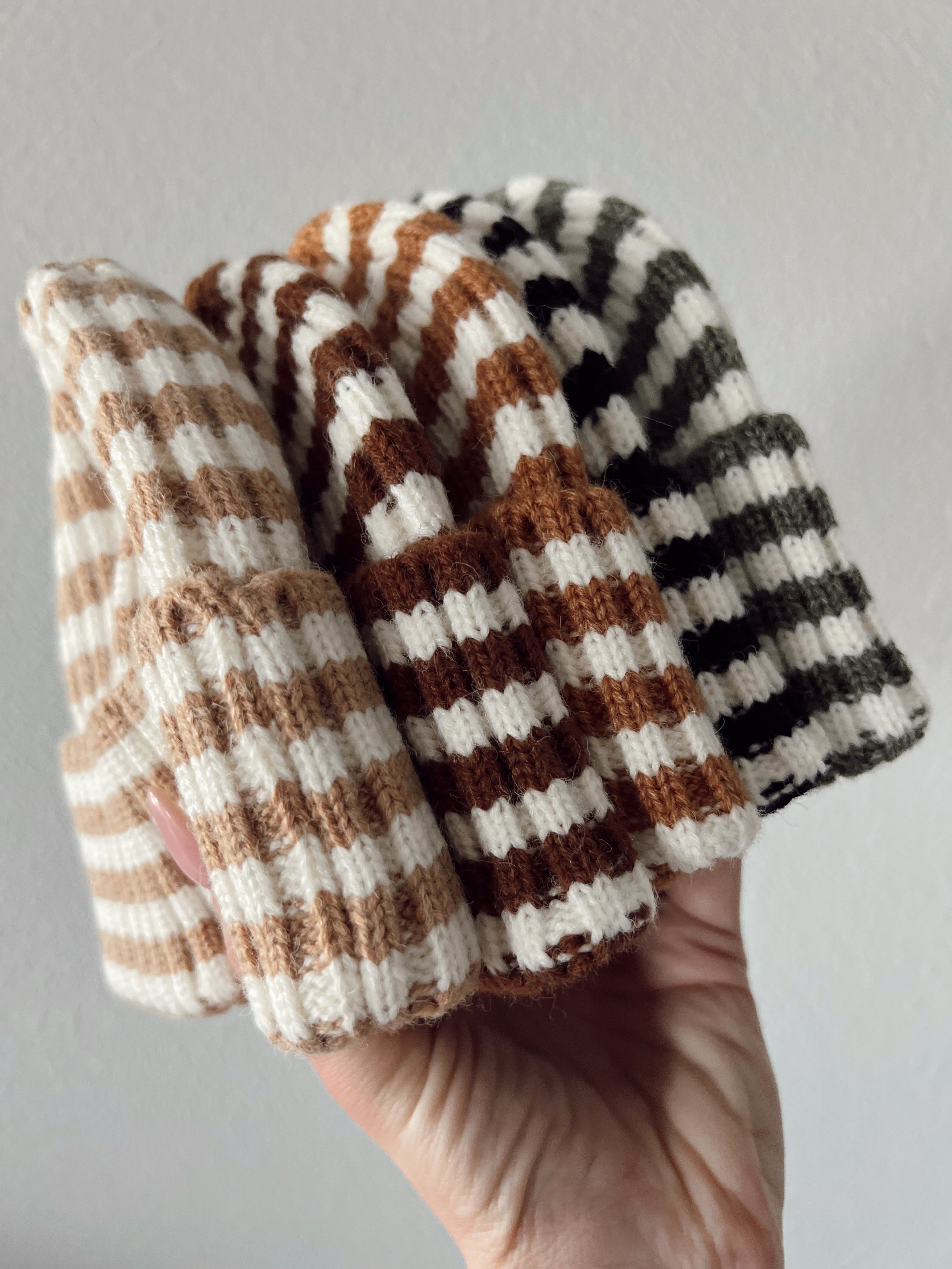 A hand holding four striped beanies in beige, brown, and green colors against a light background.