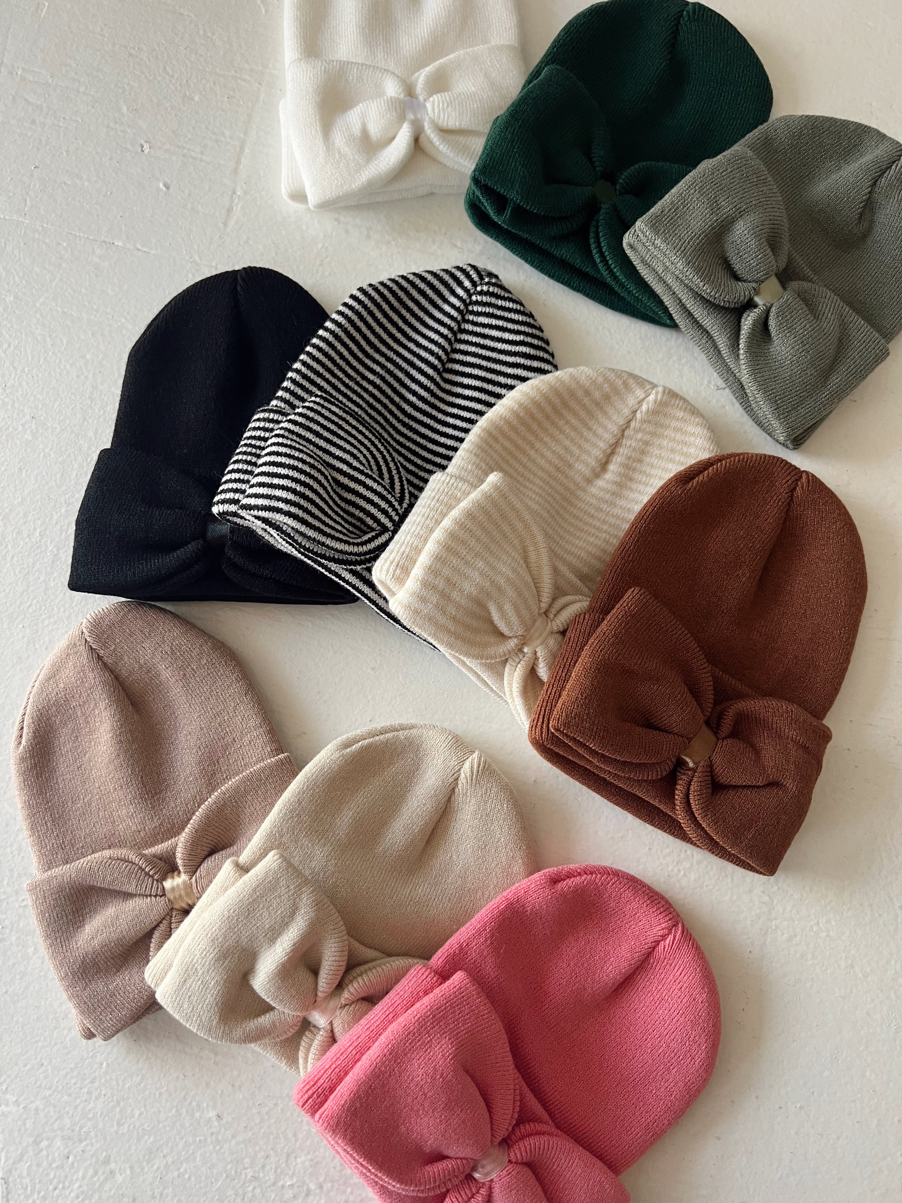 Collection of knit beanies in various colors and styles, including bow accents, arranged on a white surface.