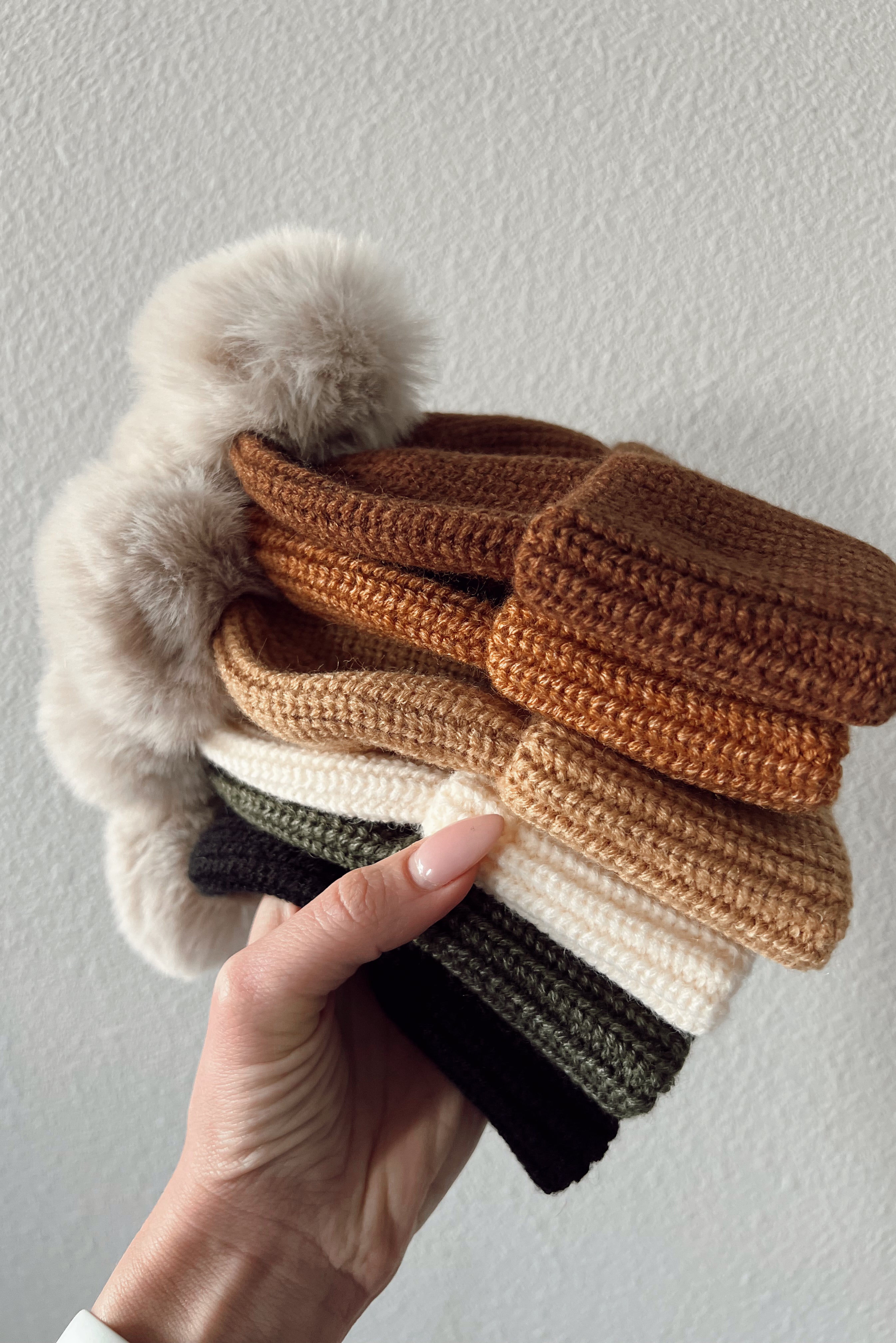 A hand holds a stack of knitted hats in earthy colors with fluffy pom-poms on top against a light background.