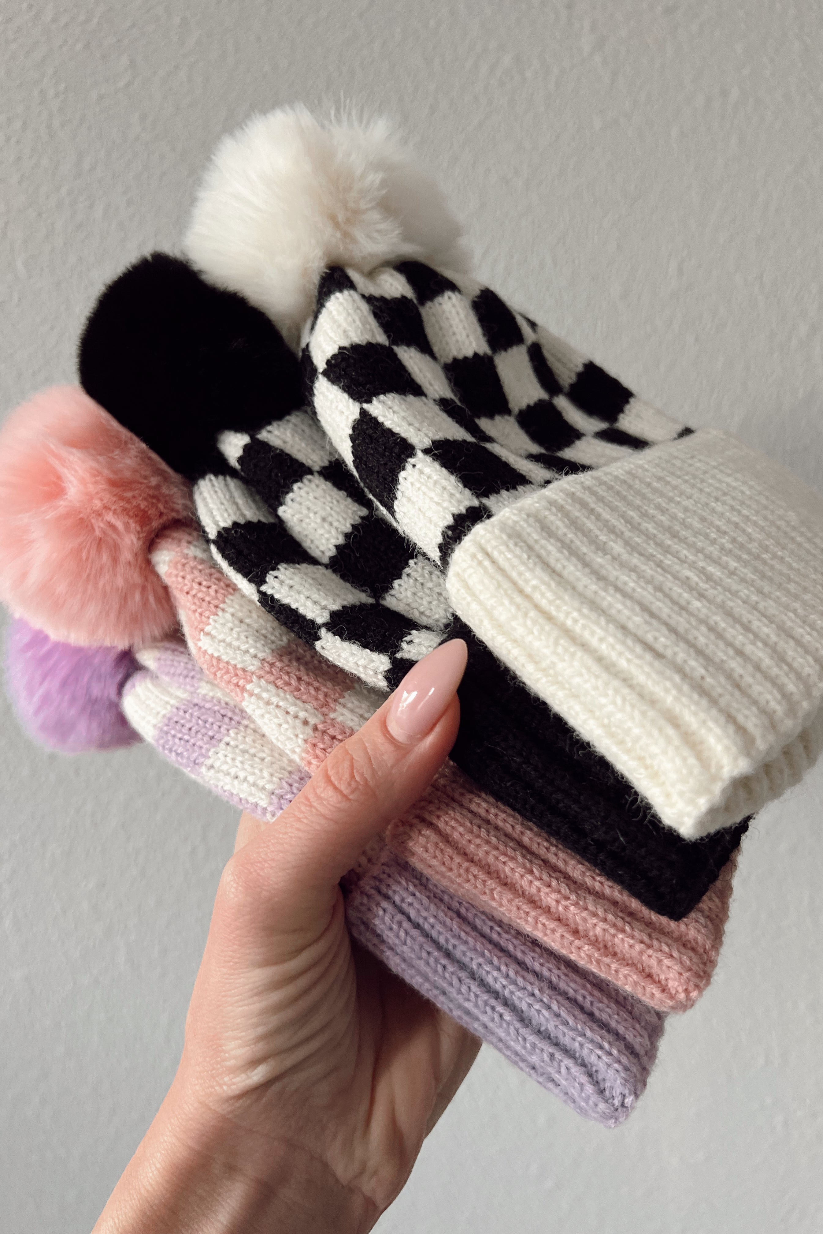 Colorful knitted hats with pom poms stacked in a hand against a neutral background.