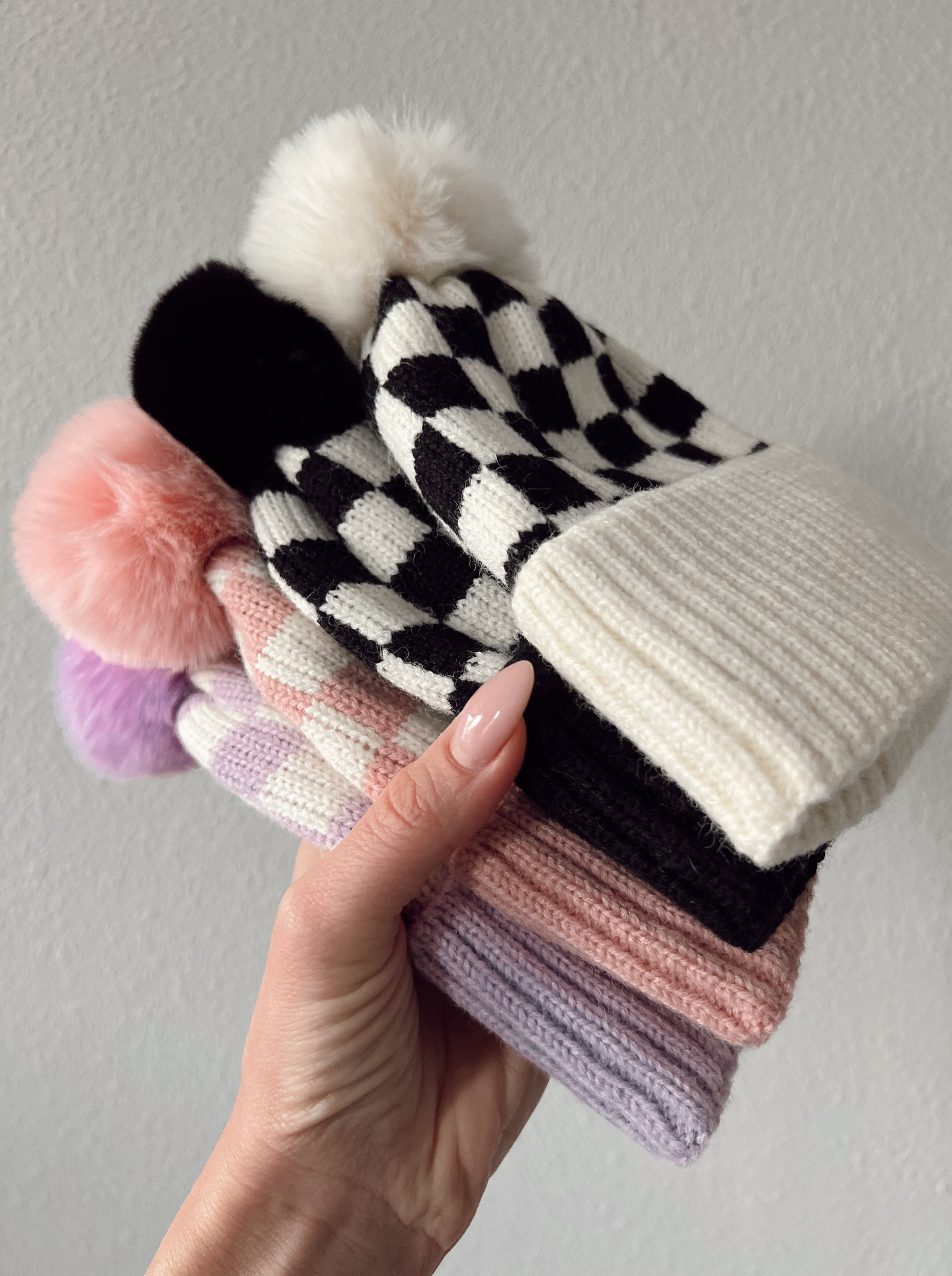 Colorful knitted hats with pom poms stacked in a hand against a neutral background.