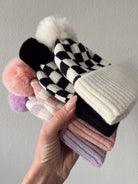 Colorful knitted hats with pom poms stacked in a hand against a neutral background.
