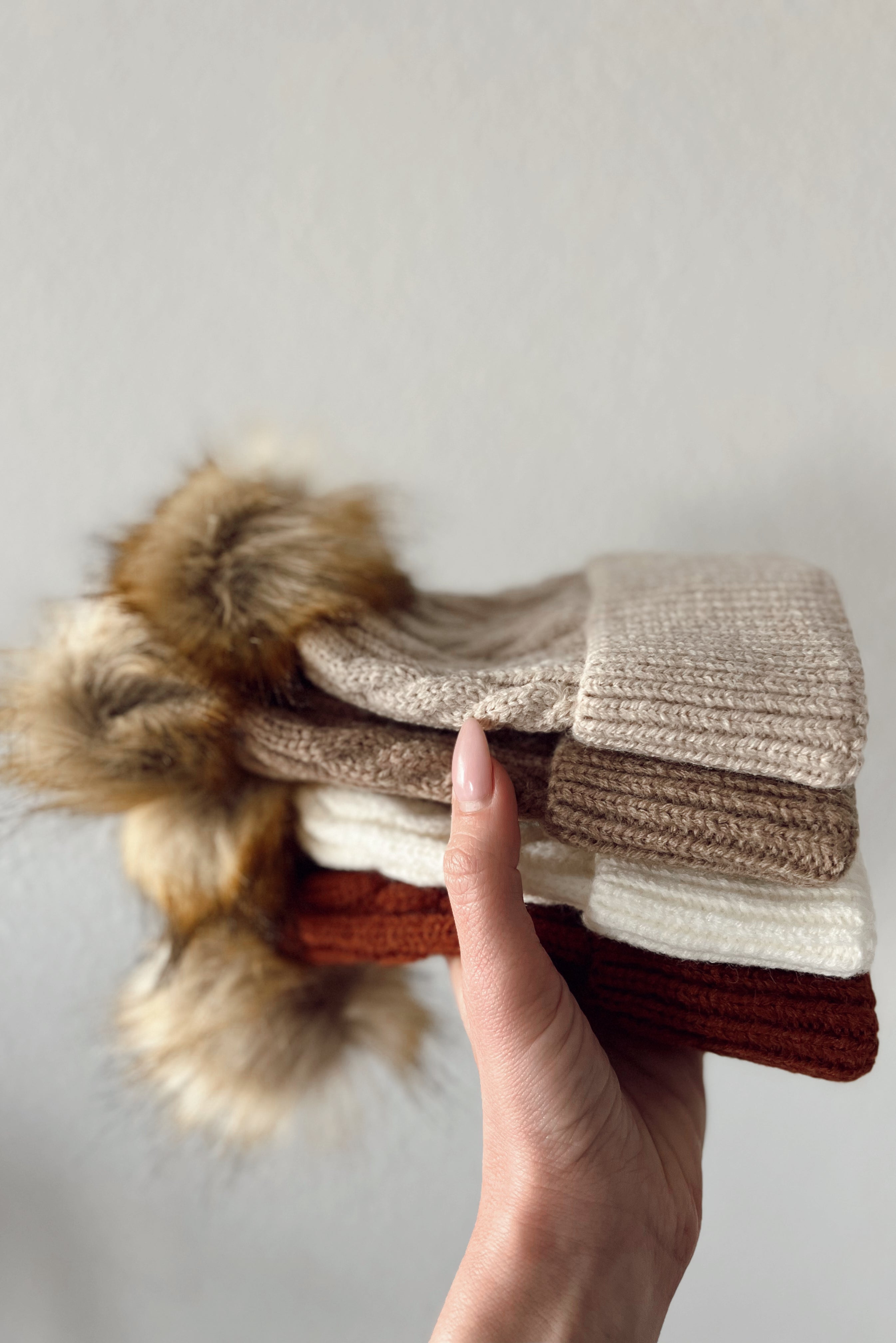 Hand holding a stack of cozy knitted hats in beige, brown, and cream, featuring fluffy pom-poms.