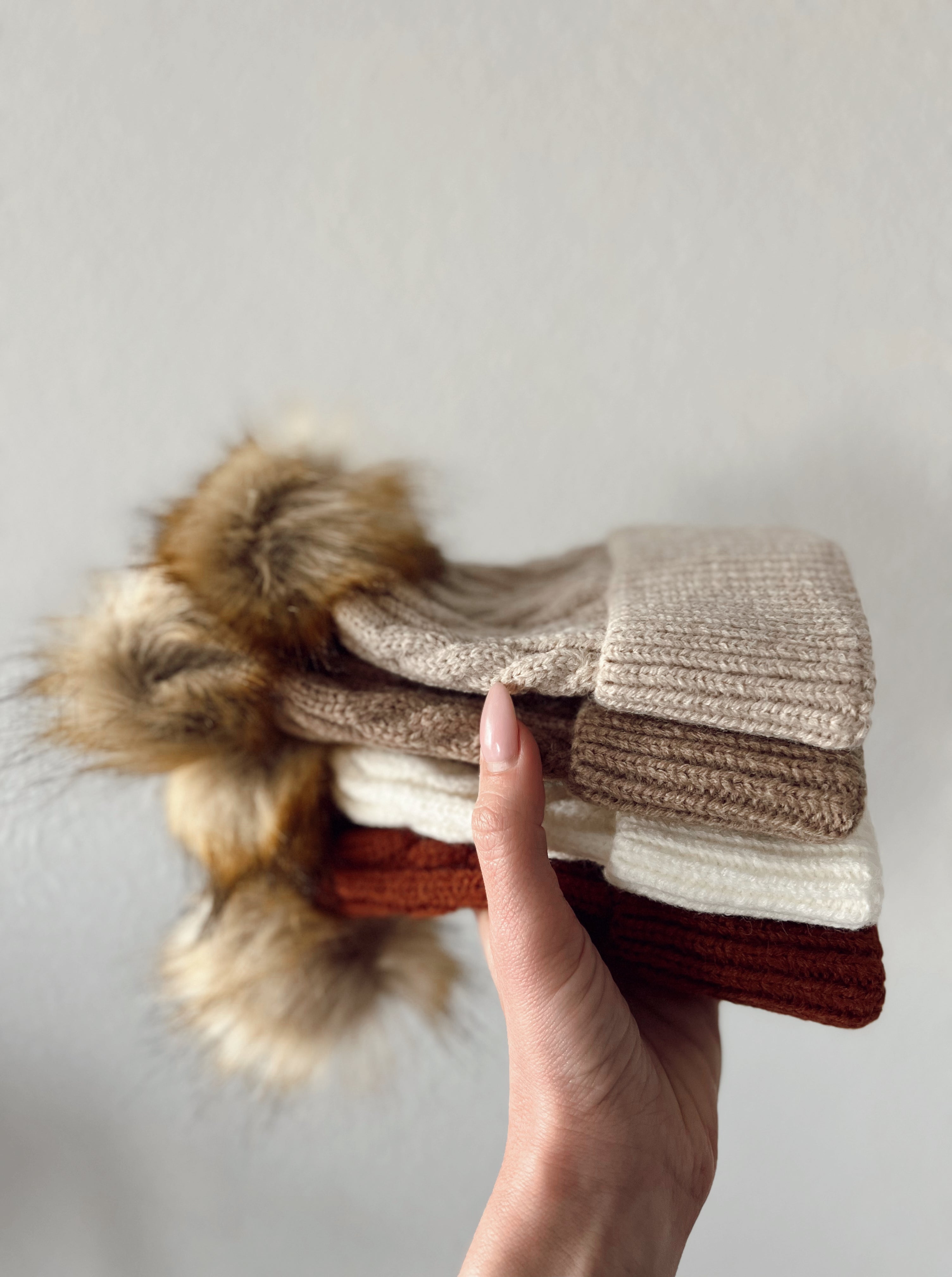 Hand holding a stack of cozy knitted hats in beige, brown, and cream, featuring fluffy pom-poms.