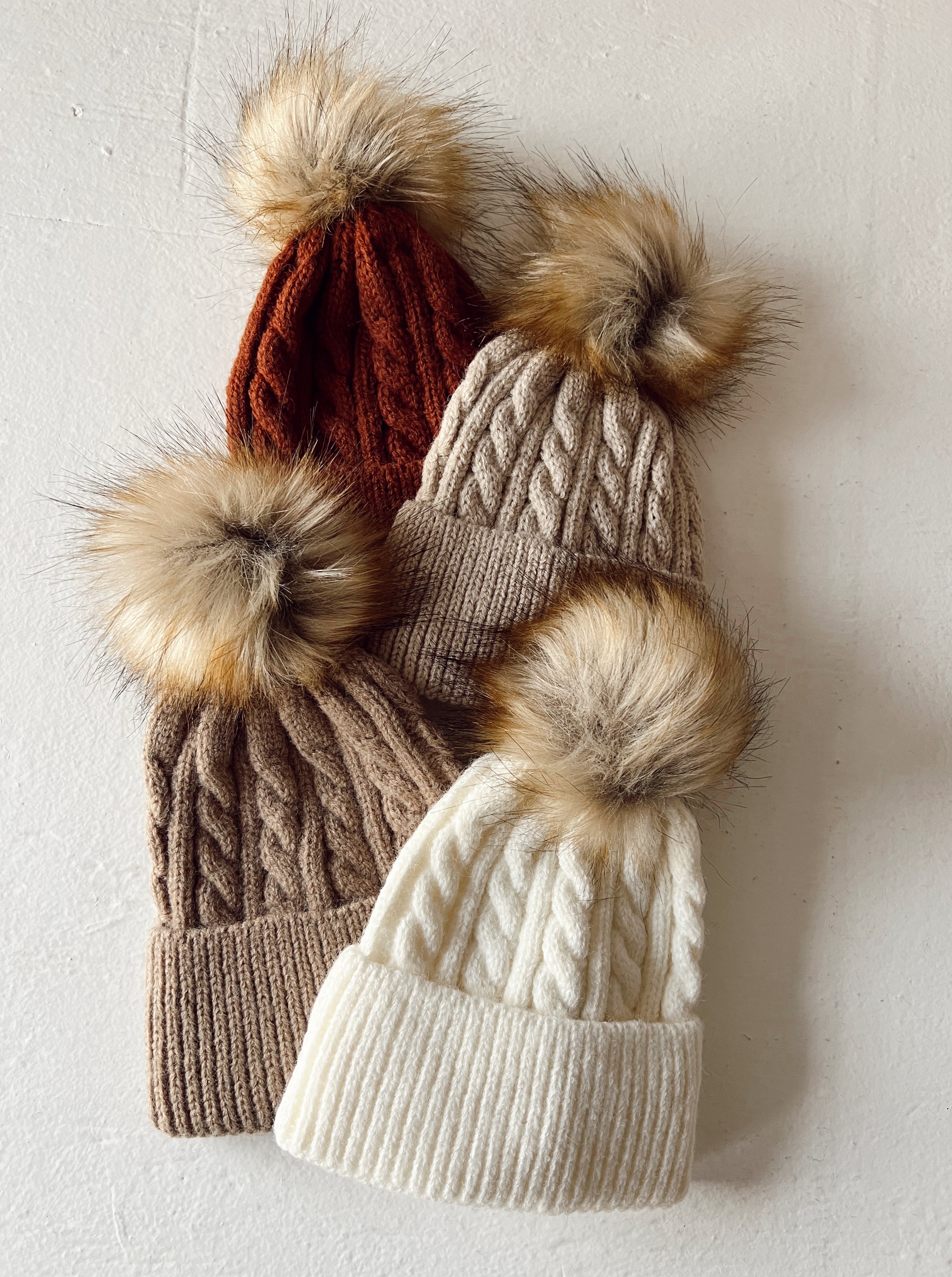 Four knitted beanies in various colors with fur pom-poms on top, arranged on a light background.