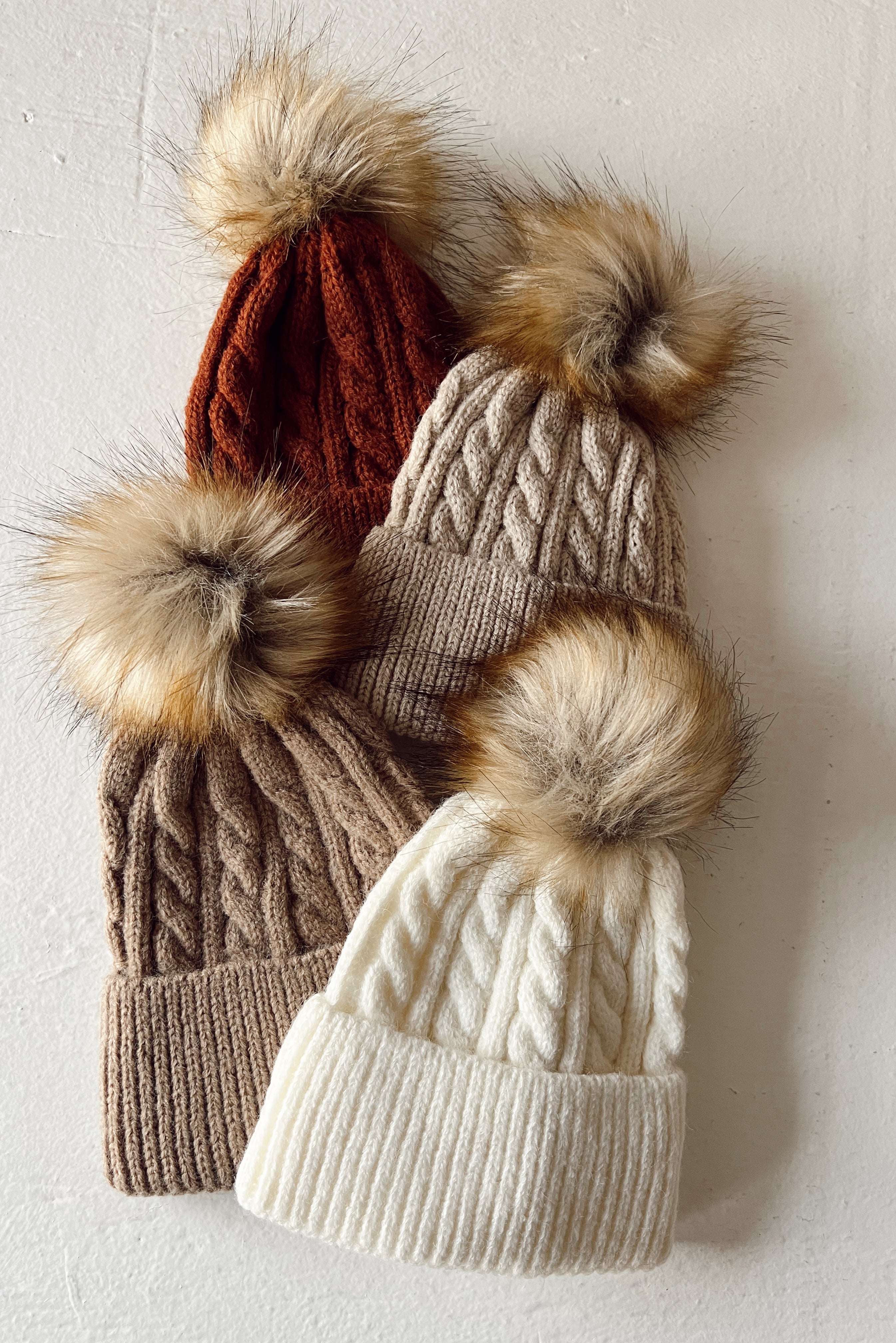 Four knitted beanies in earthy tones with fur pom-poms, arranged on a light background.