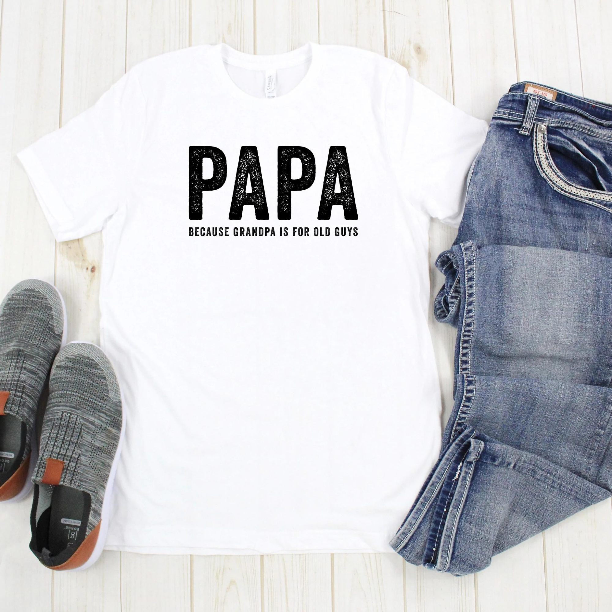 White t-shirt with "PAPA" text and "because grandpa is for old guys," paired with jeans and gray shoes on a wooden background.