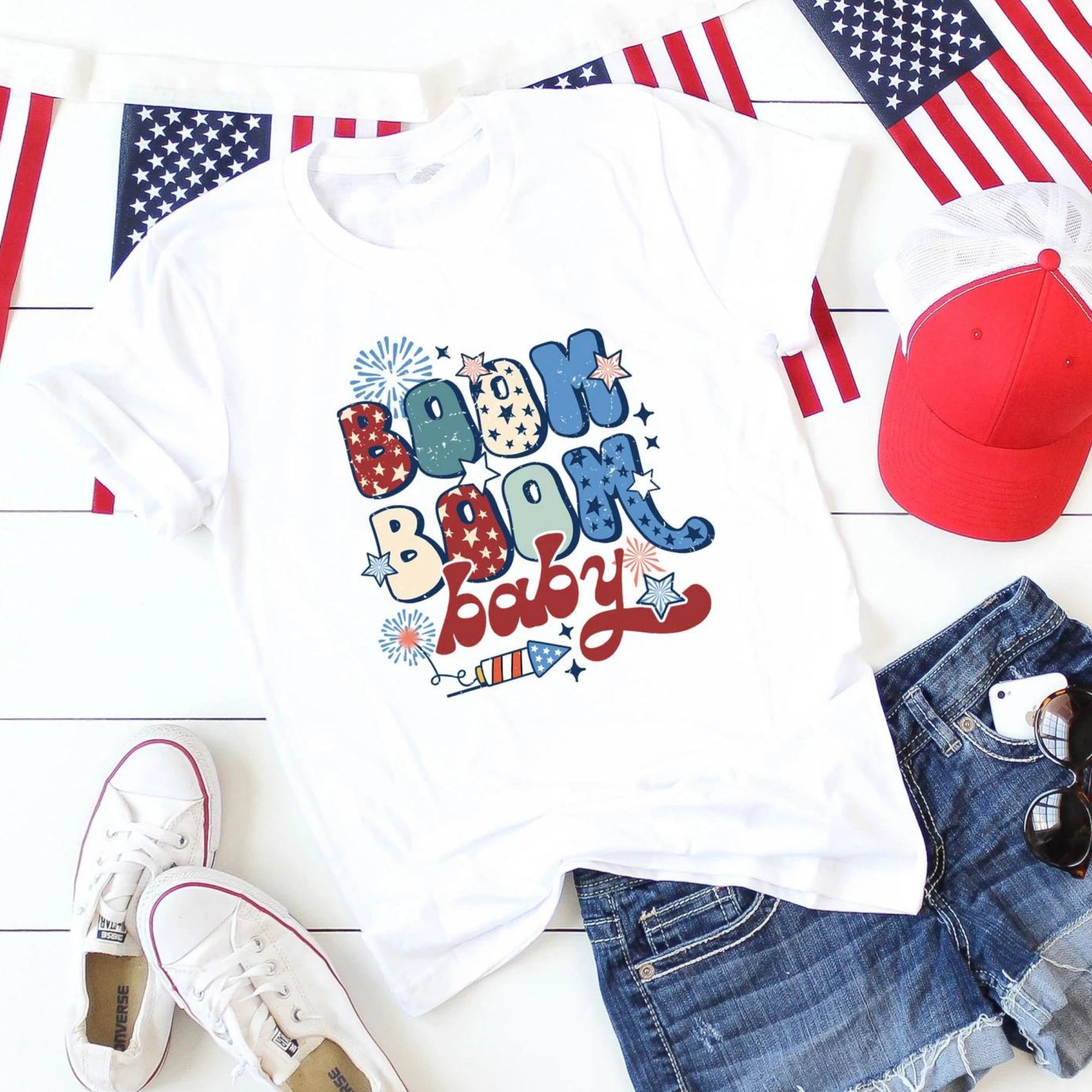 White t-shirt with "Boom Boom Baby" design, surrounded by denim shorts, sneakers, red cap, and American flags.