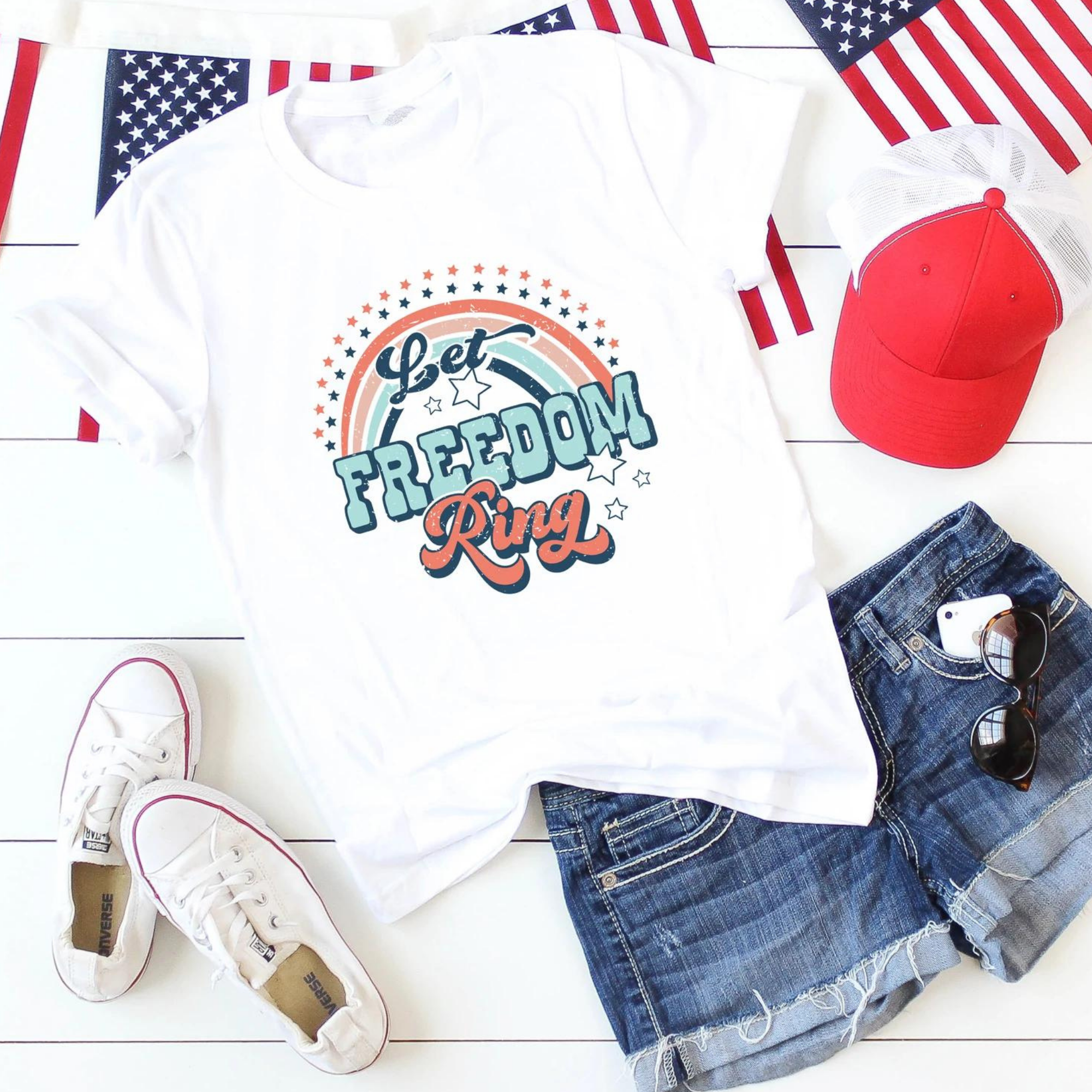 White T-shirt with "Let Freedom Ring" design, paired with denim shorts, red cap, sneakers, and sunglasses.