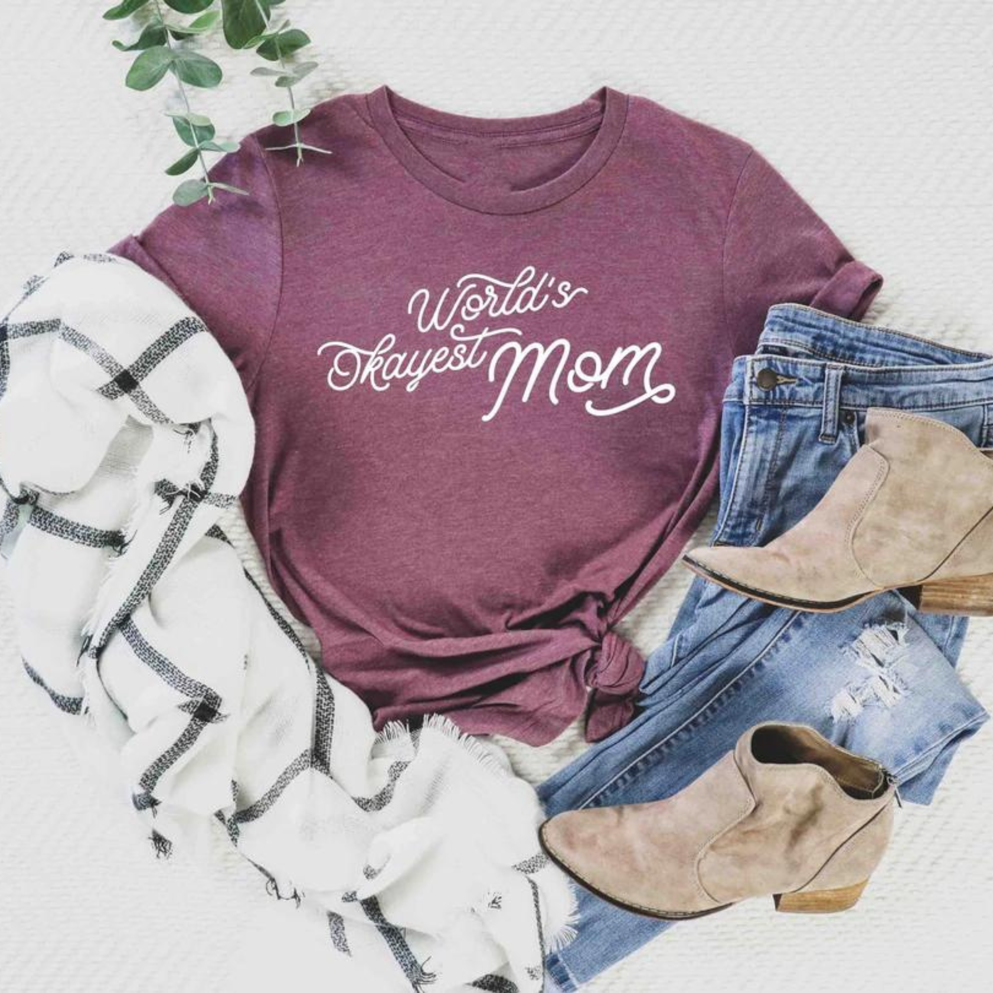 Maroon t-shirt with "World's Okayest Mom" text, styled with jeans, taupe boots, and a white plaid scarf.