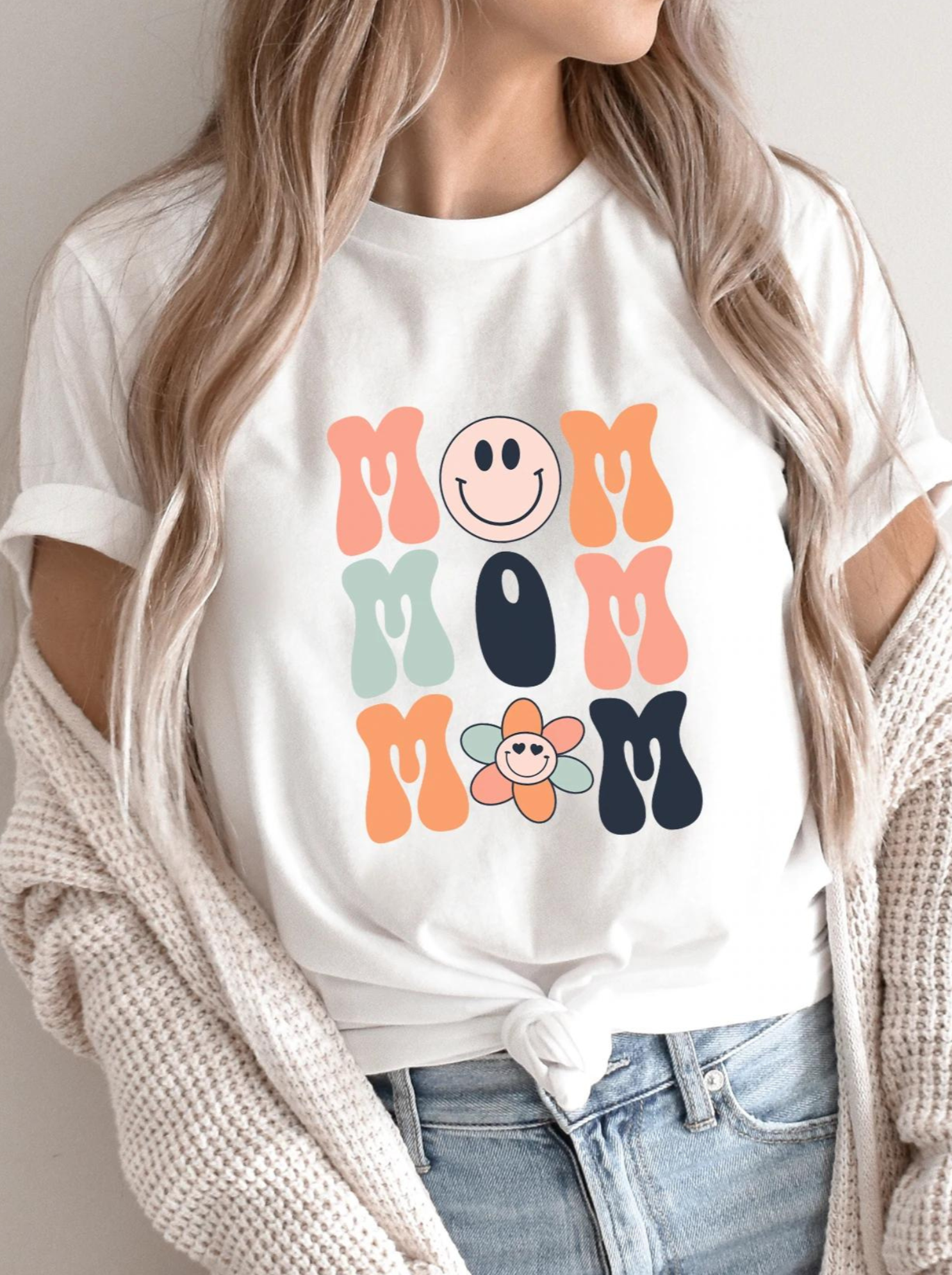 Woman wearing a white t-shirt with colorful "MOM" text and a smiley flower graphic.
