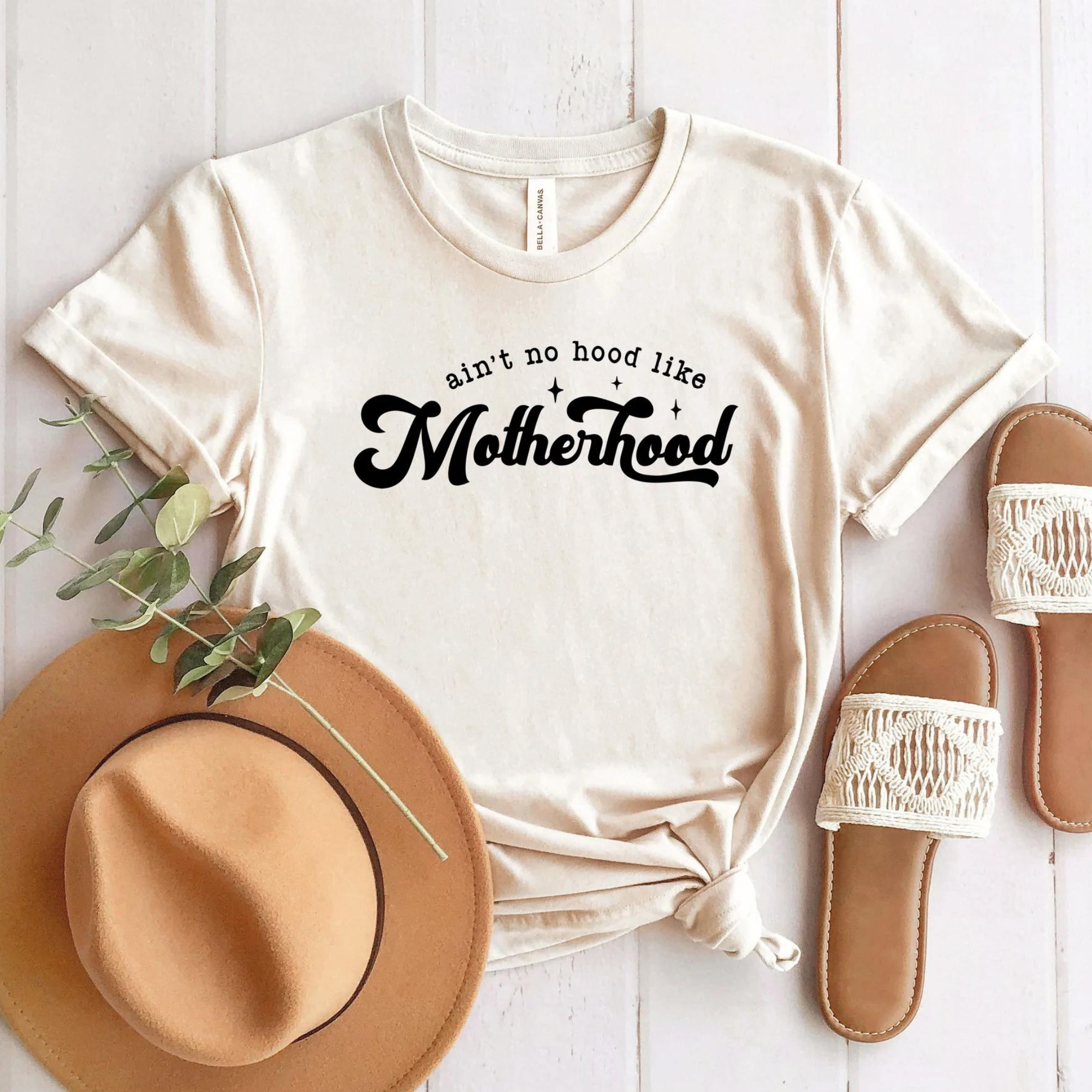 Beige t-shirt with "ain't no hood like Motherhood" text, a hat, and sandals on a white wooden surface.