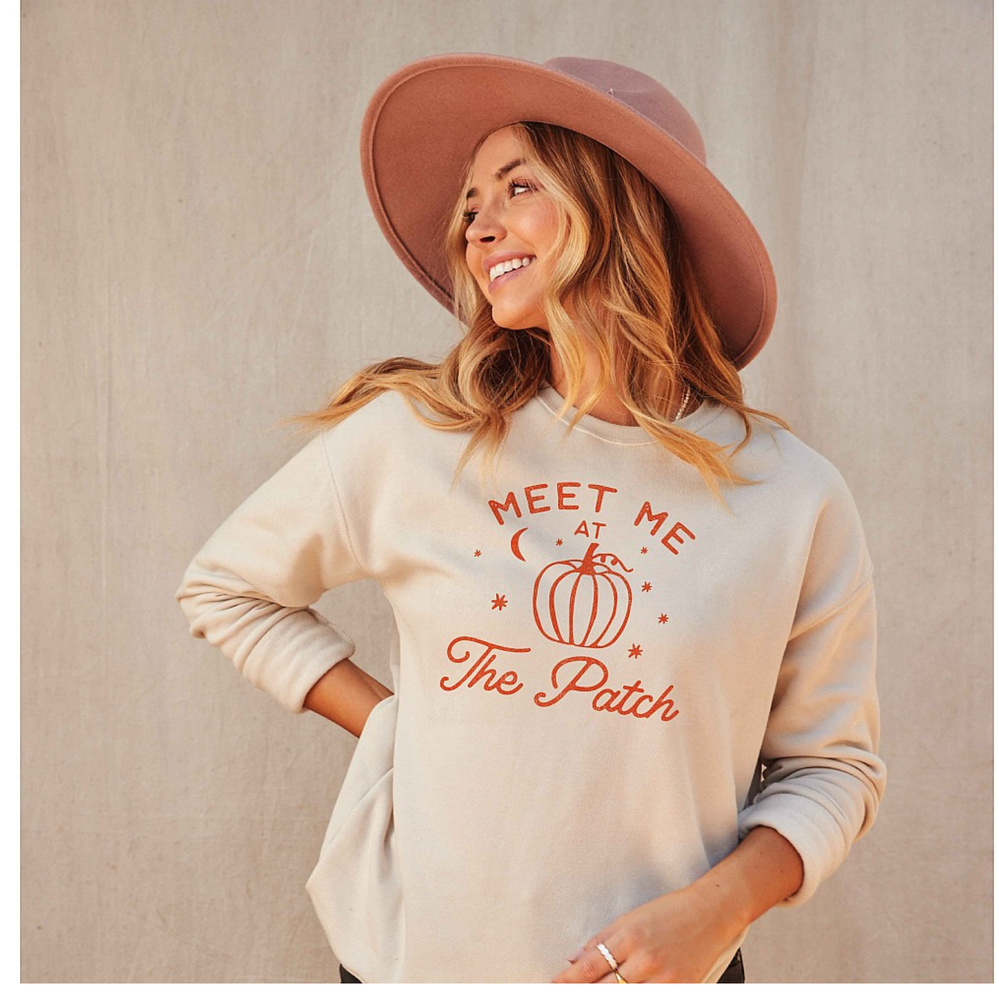 Woman smiling in a beige sweatshirt with a pumpkin graphic, wearing a pink hat against a neutral background.