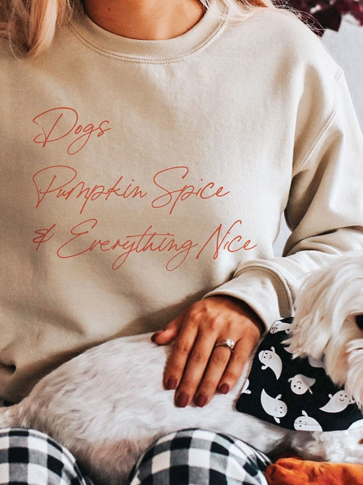 A woman in a cozy sweatshirt pets a small dog with a ghost-patterned bandana, sitting comfortably indoors.