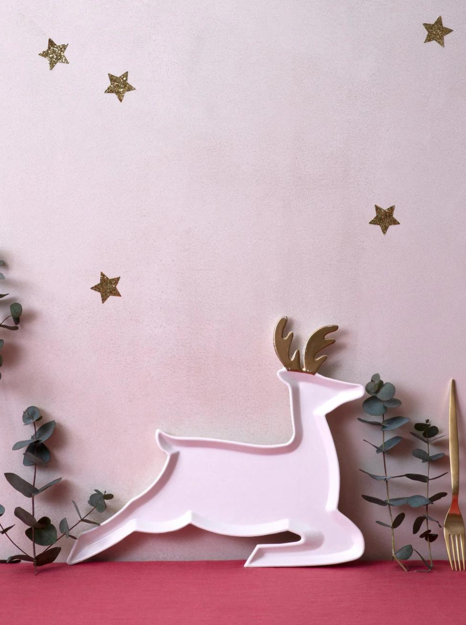 White reindeer platter next to green eucalyptus leaves and golden cutlery on a pink background with gold stars.