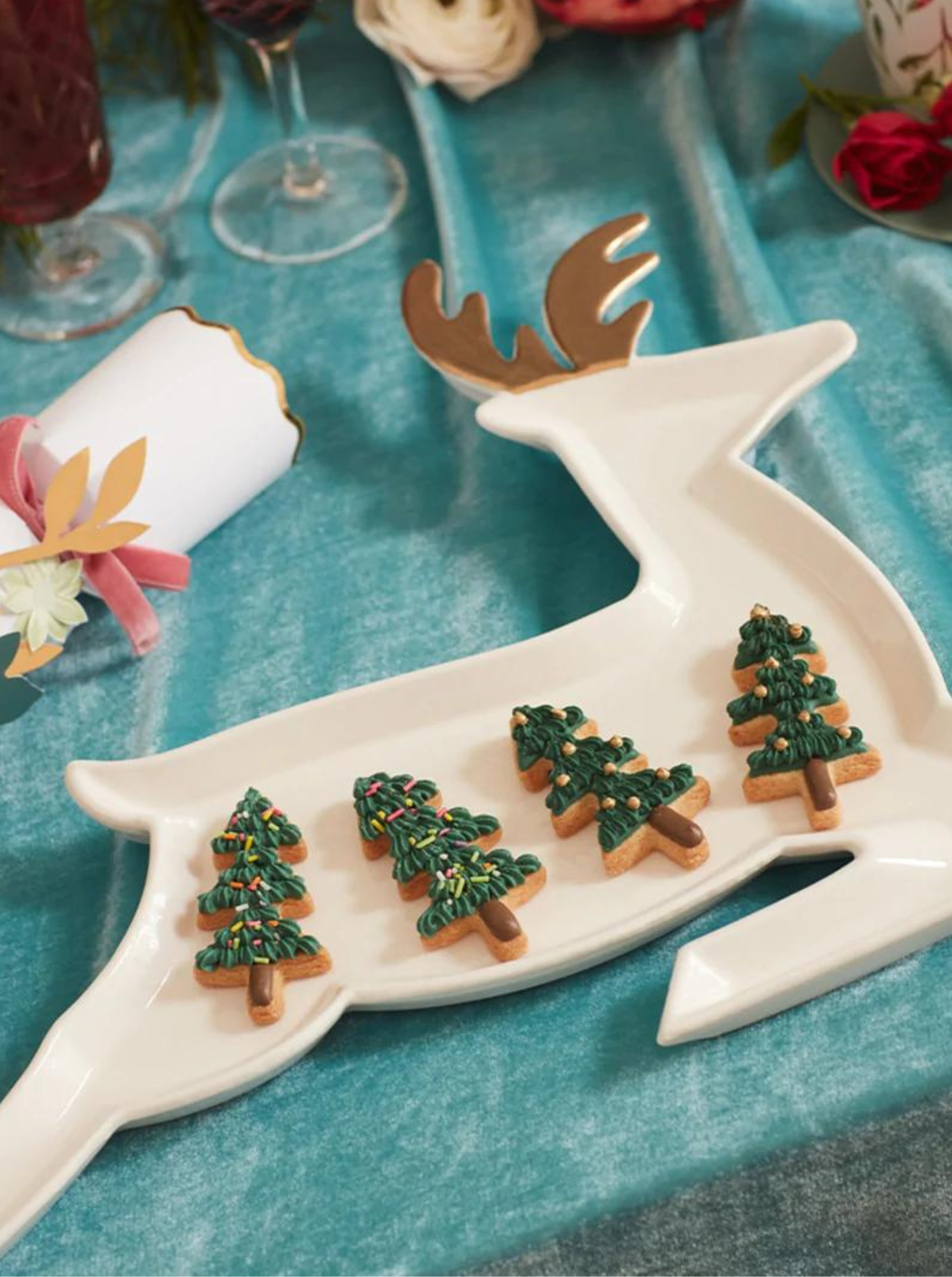 White reindeer platter with decorated tree-shaped cookies, set on a teal tablecloth with festive decorations.