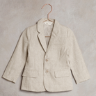Light beige children's blazer with two buttons, hanging on a copper hook against a textured background.