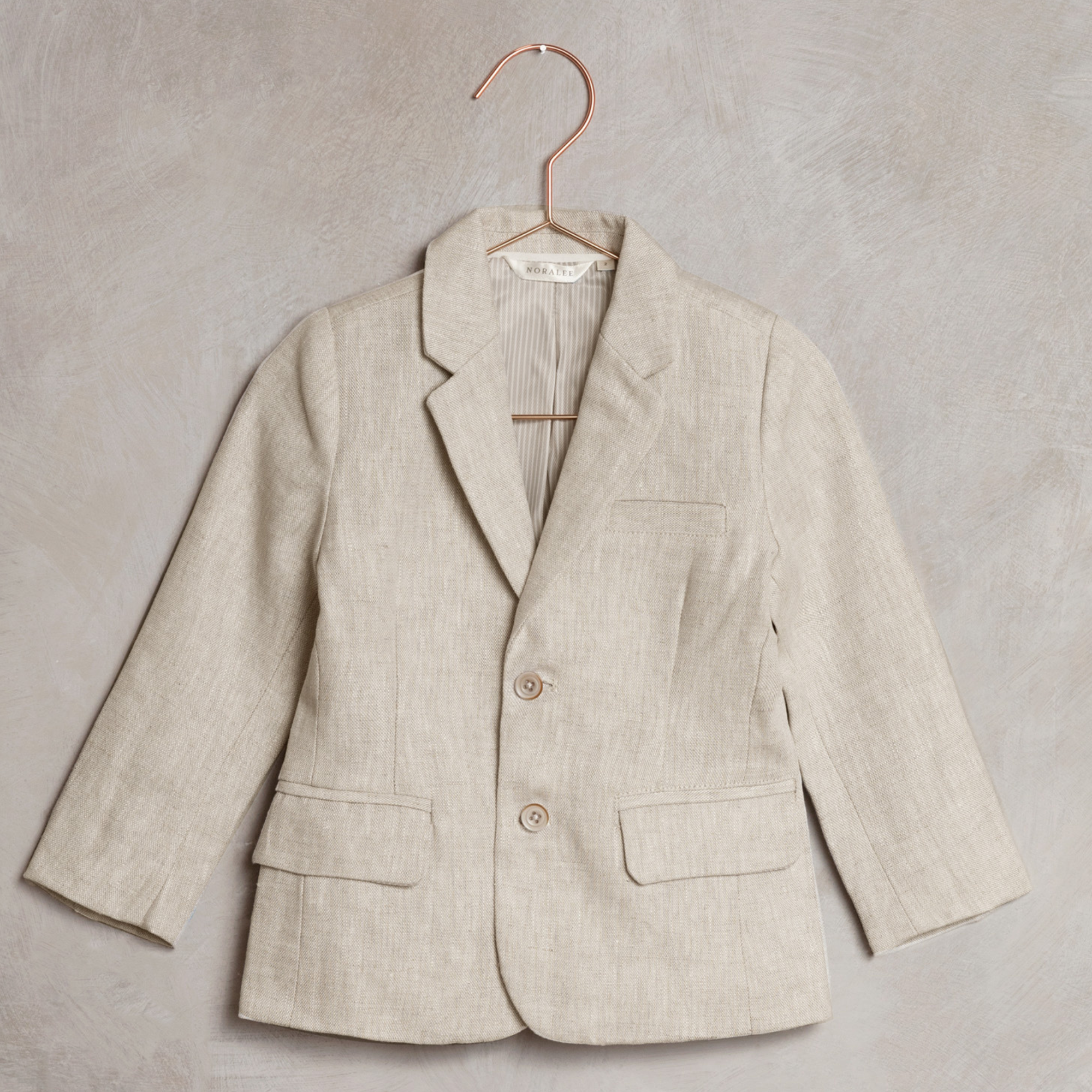 Light beige children's blazer with two buttons, hanging on a copper hook against a textured background.