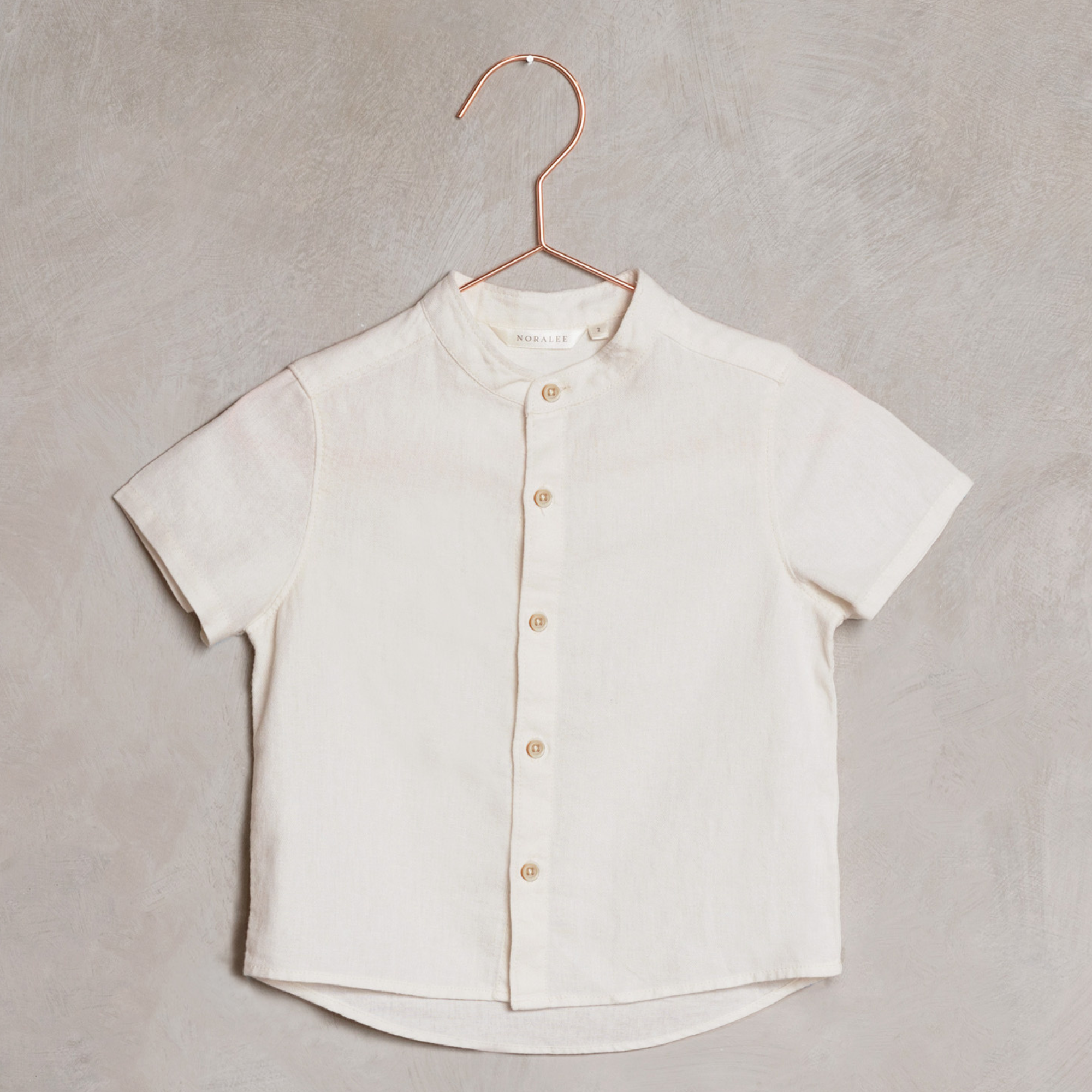 Short-sleeve, beige collared shirt hanging on a copper hook against a textured gray background.