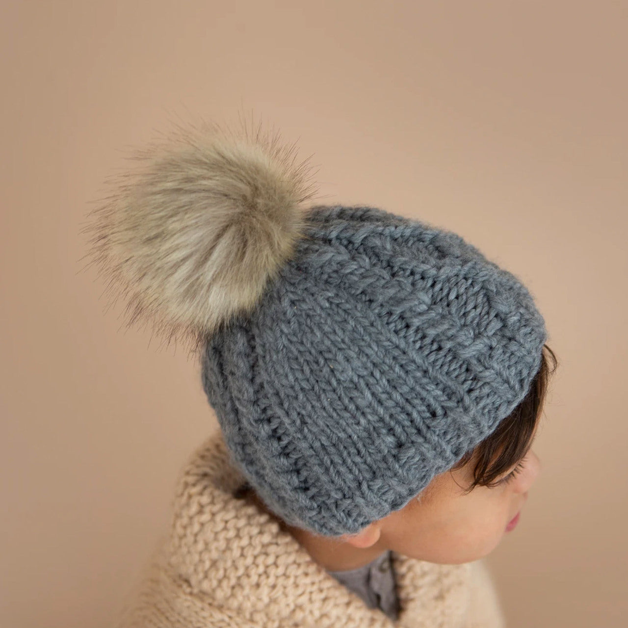 Child wearing a gray knitted hat with a fluffy pom-pom against a soft beige background.