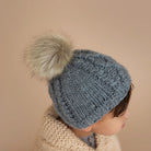 Child wearing a gray knitted hat with a fluffy pom-pom against a soft beige background.