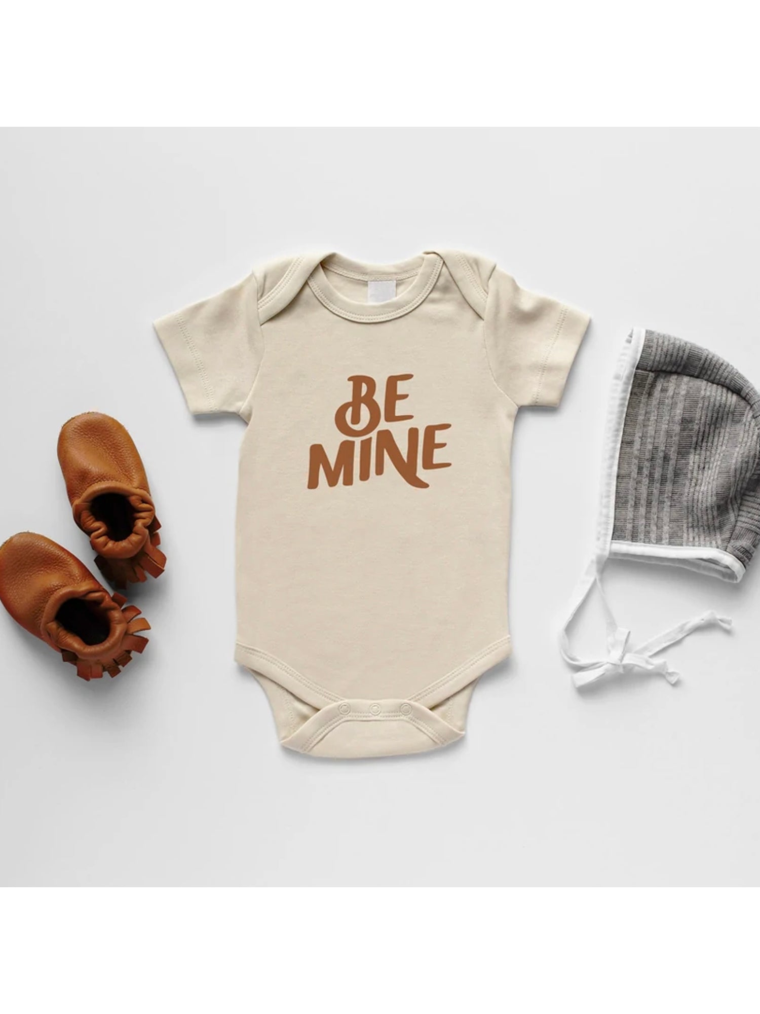 Beige baby onesie with "BE MINE" text, brown shoes, and a gray striped hat, styled on a white background.