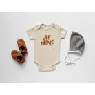 Beige baby onesie with "BE MINE" text, brown shoes, and a gray striped hat, styled on a white background.
