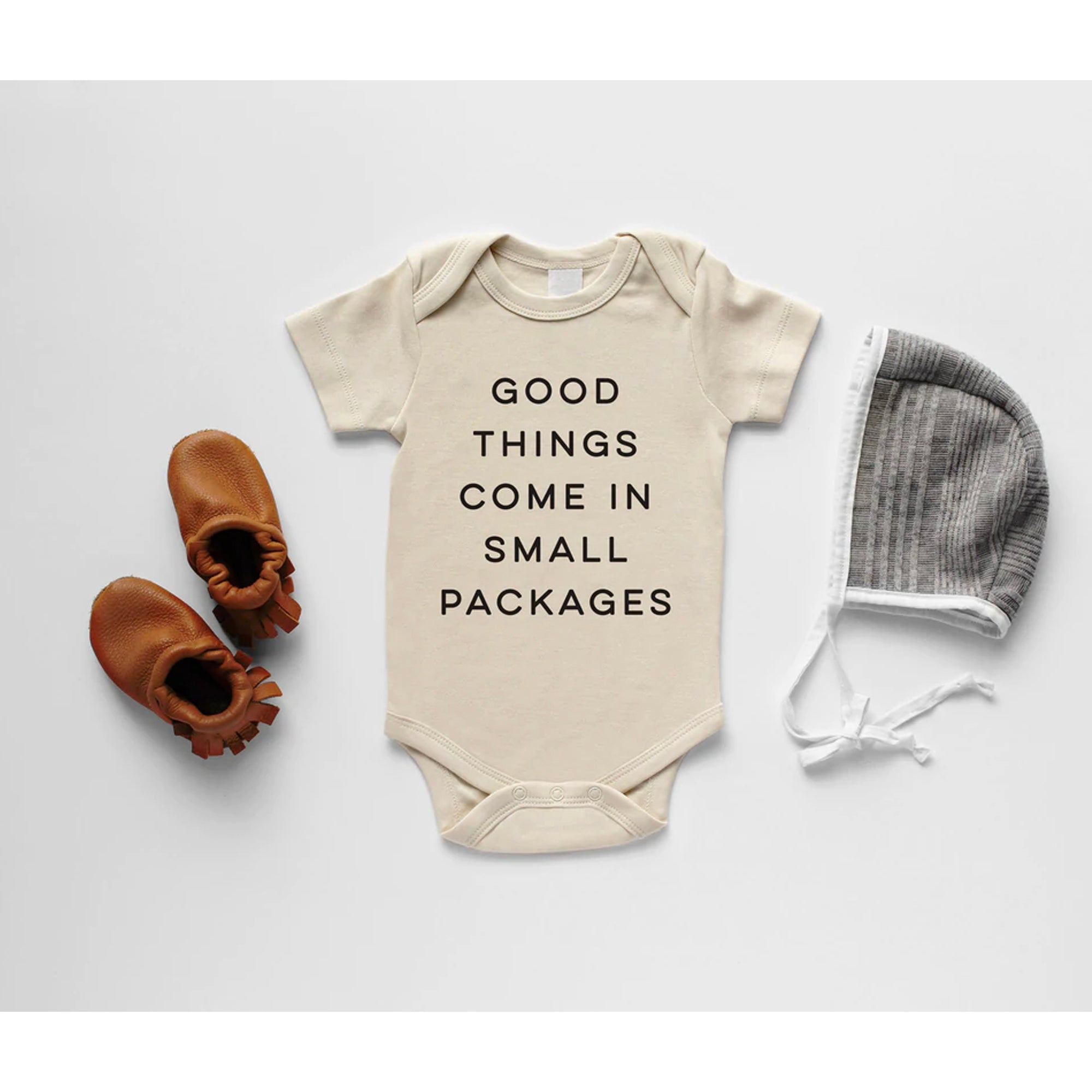 Beige baby onesie with "Good things come in small packages" text, paired with brown booties and a gray baby hat.