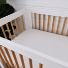 White crib with natural wooden slats and a soft, padded mattress, set against a light background and green plant.