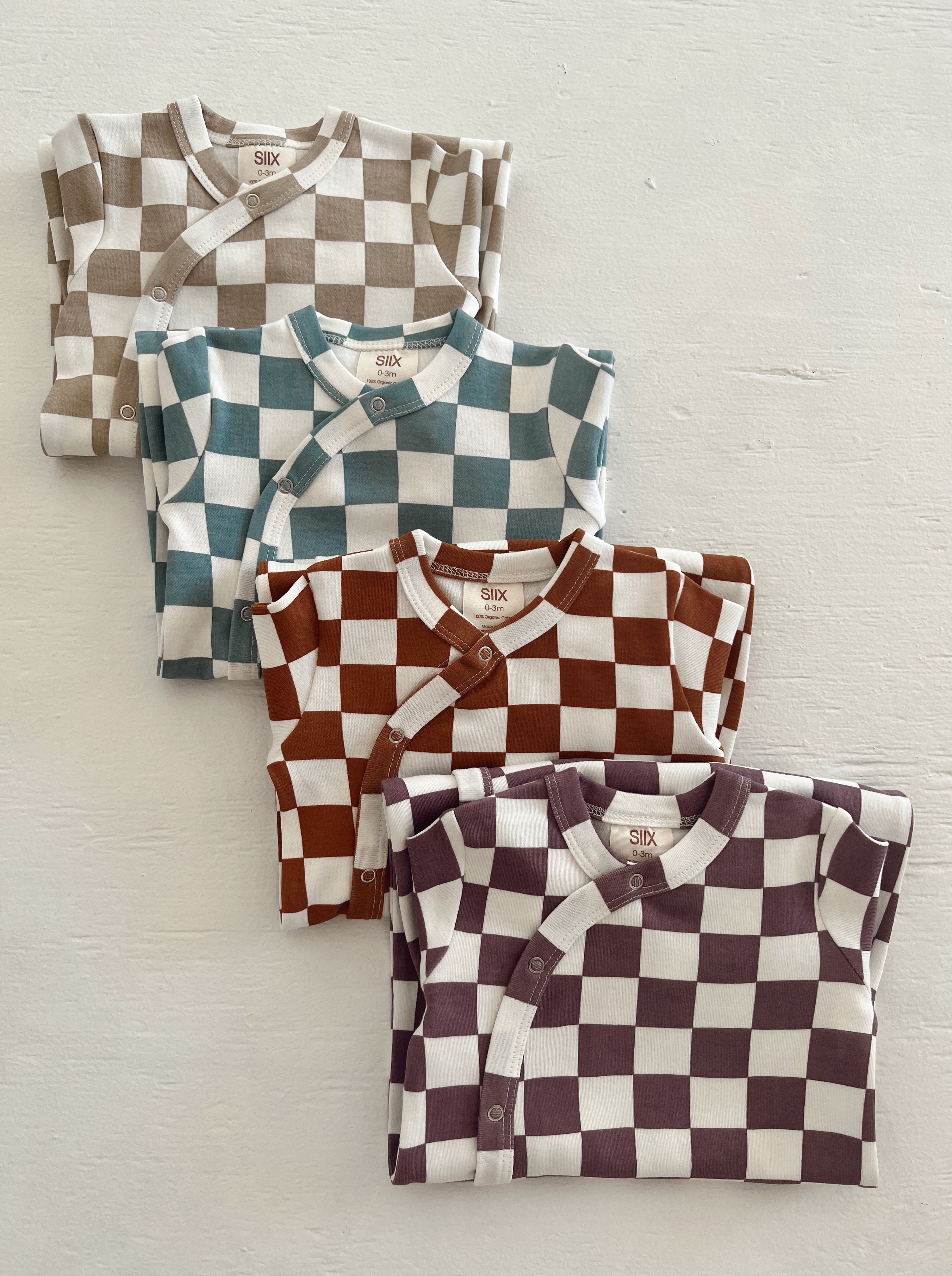 Four checkered baby onesies in brown, teal, maroon, and beige, neatly stacked on a light background.