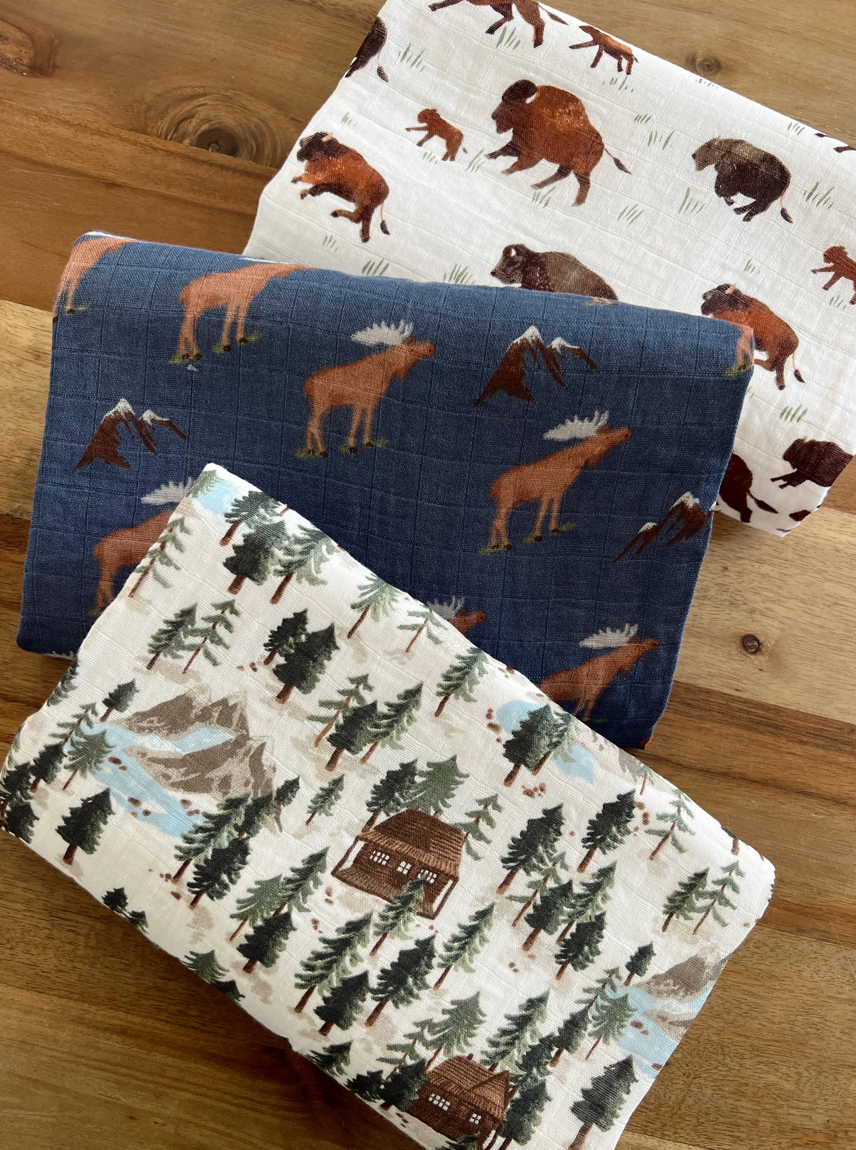 Three patterned nature-themed fabric blankets featuring moose, bison, and forest scenes on a wooden surface.