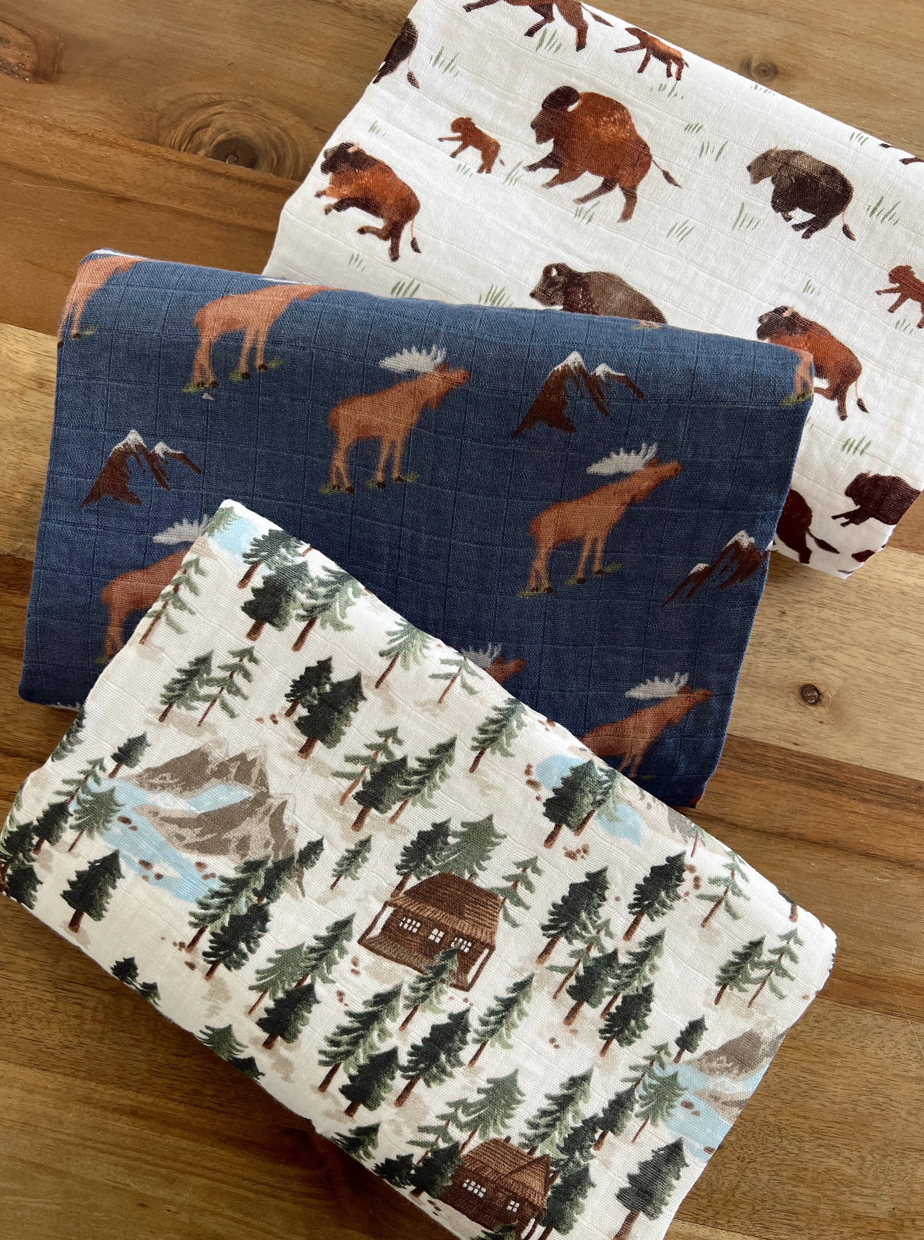 Three patterned nature-themed fabric blankets featuring moose, bison, and forest scenes on a wooden surface.