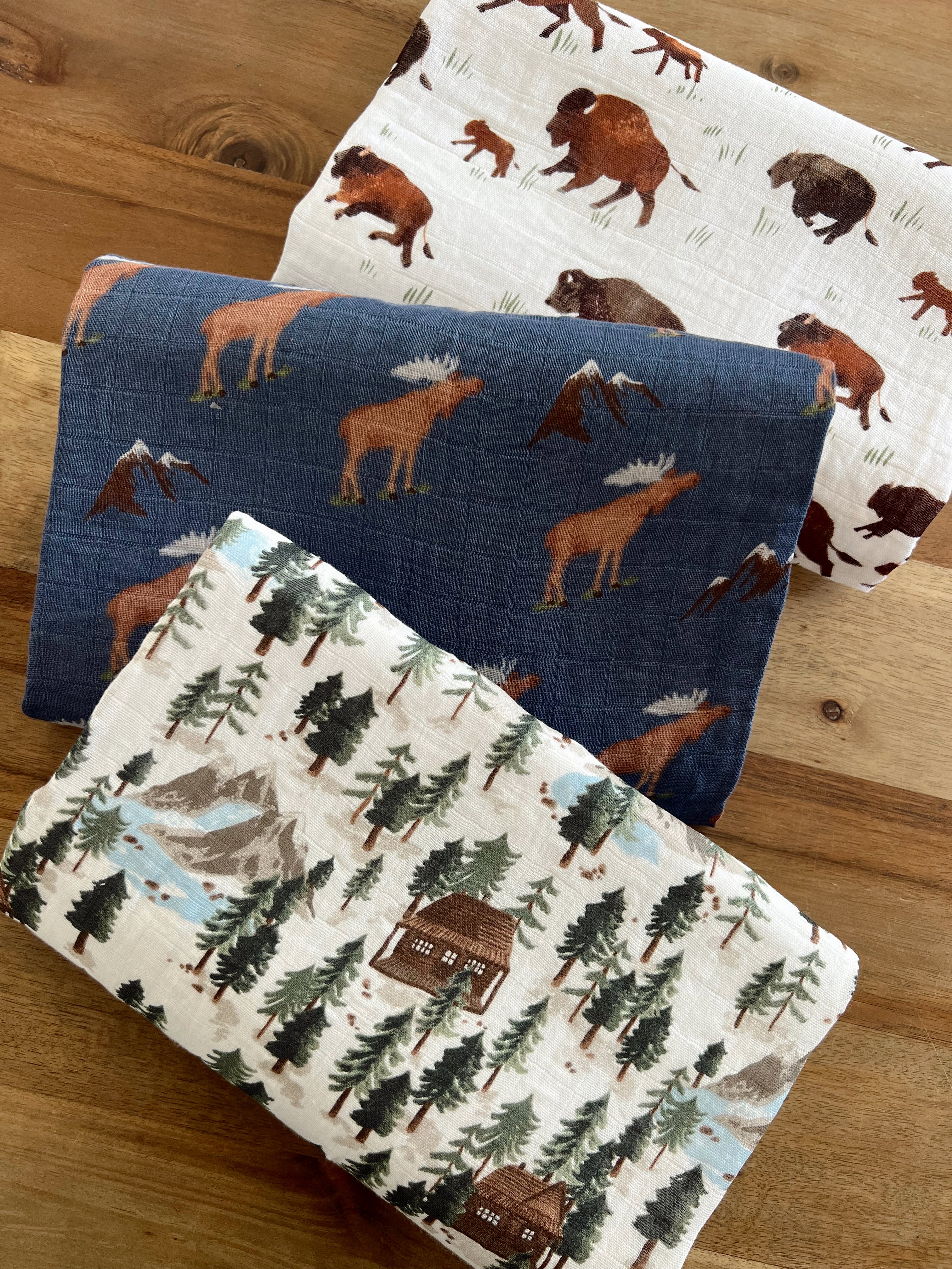 Three patterned nature-themed fabric blankets featuring moose, bison, and forest scenes on a wooden surface.