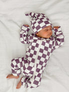 Newborn in a purple and white checkered outfit and hat, sleeping peacefully on a white blanket.