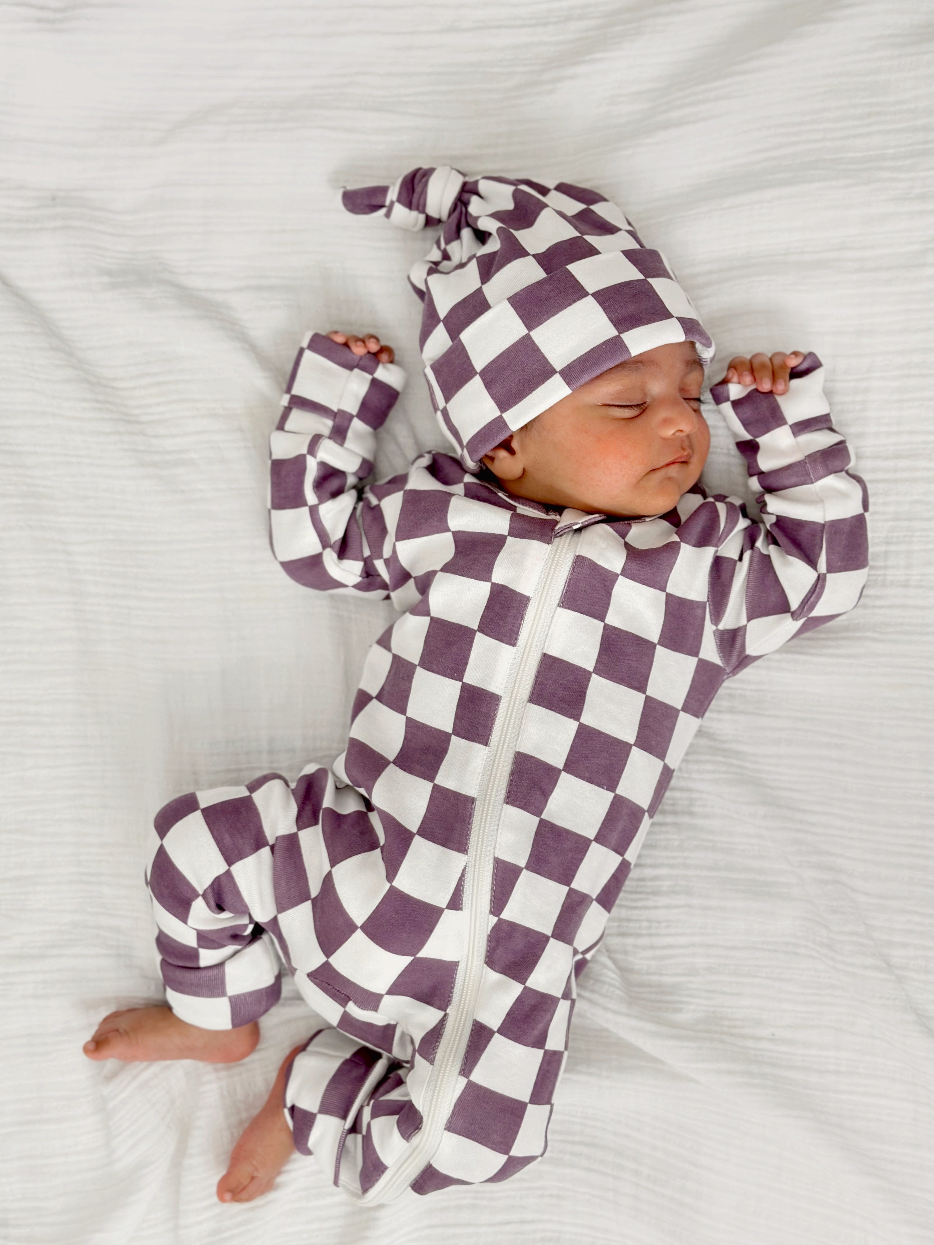 Newborn in a purple and white checkered outfit and hat, sleeping peacefully on a white blanket.