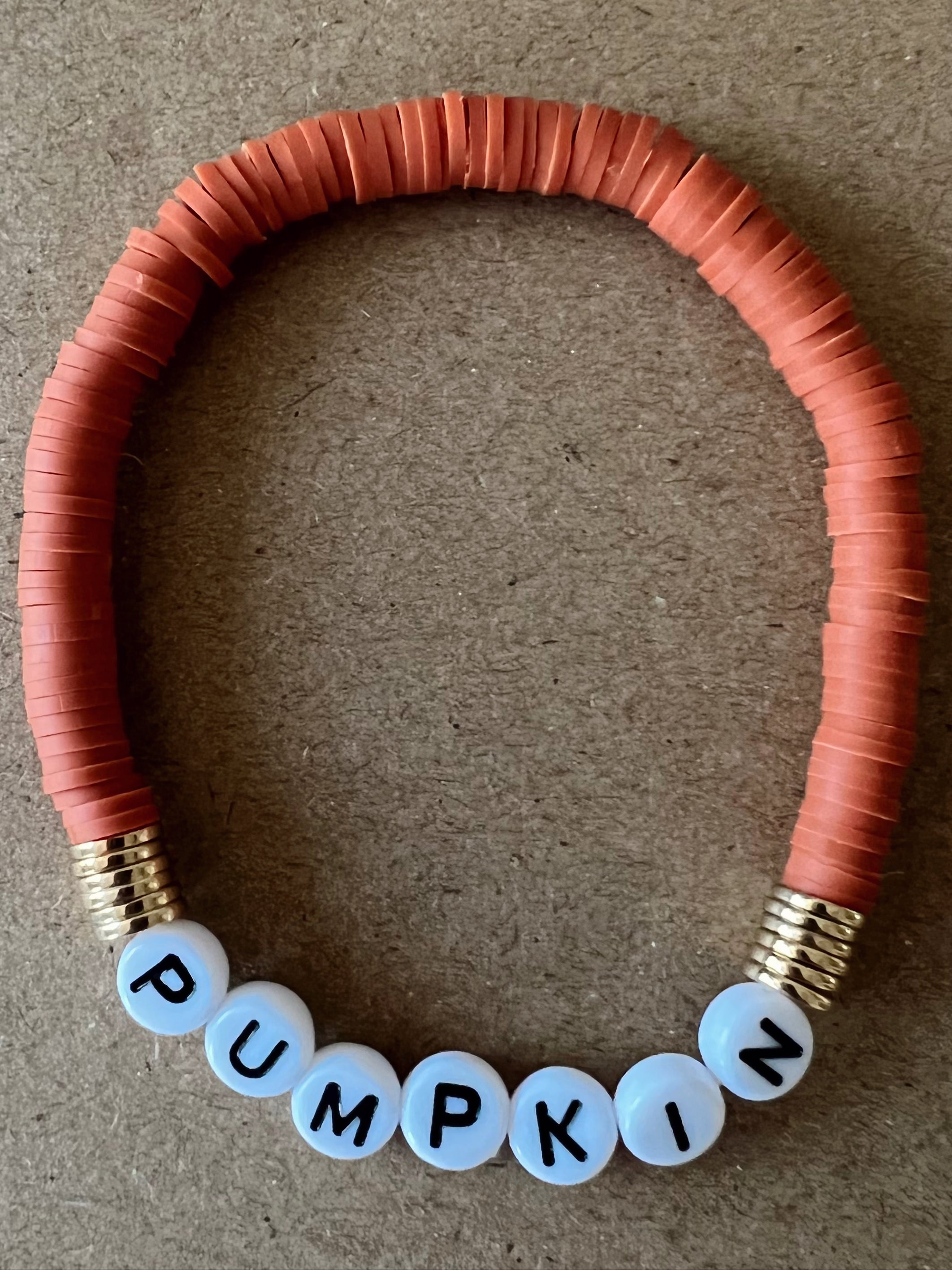 Orange beaded bracelet with white letter beads spelling "PUMPKIN" on a brown background.