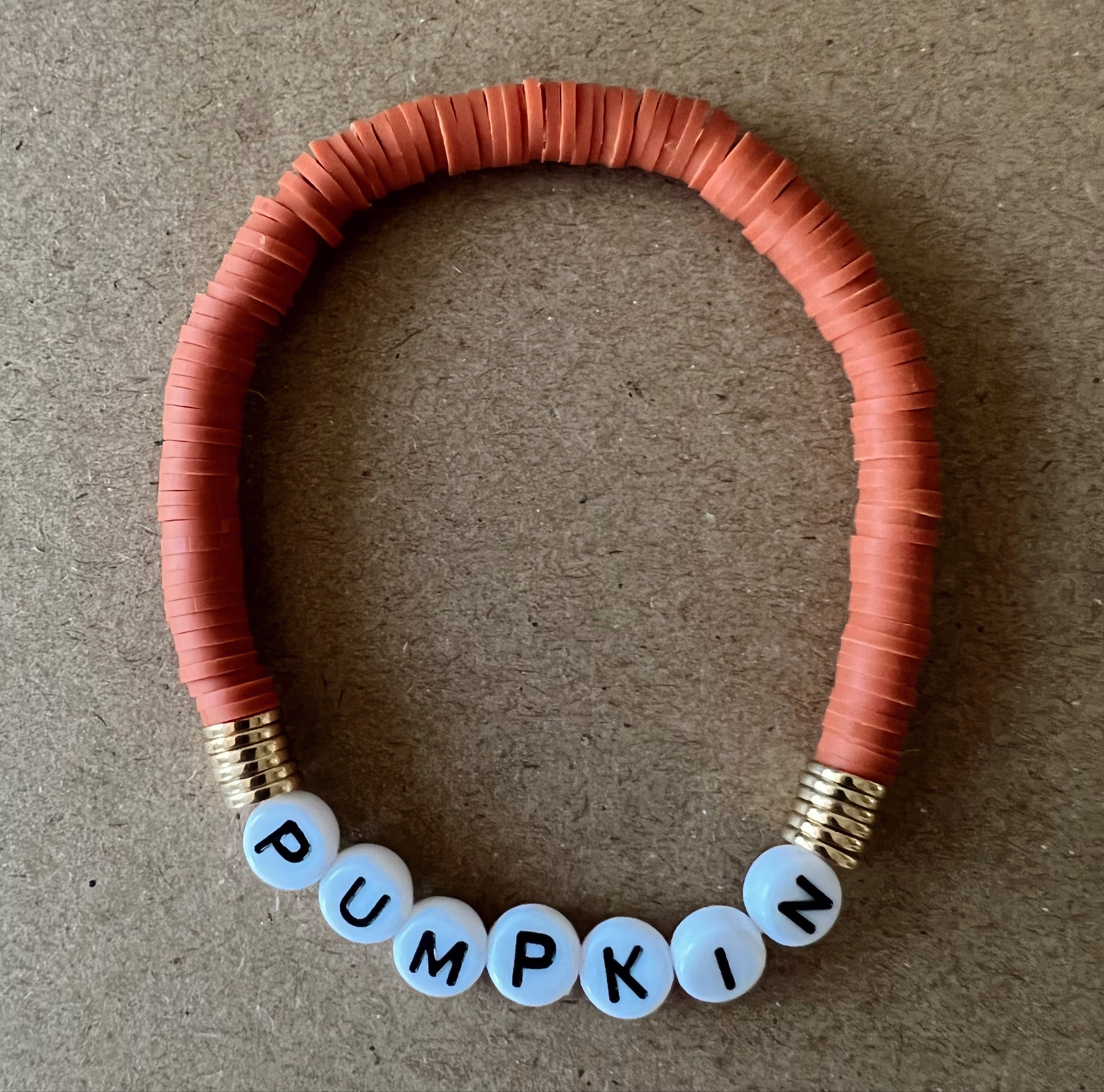 Orange beaded bracelet with white letter beads spelling "PUMPKIN" on a brown background.