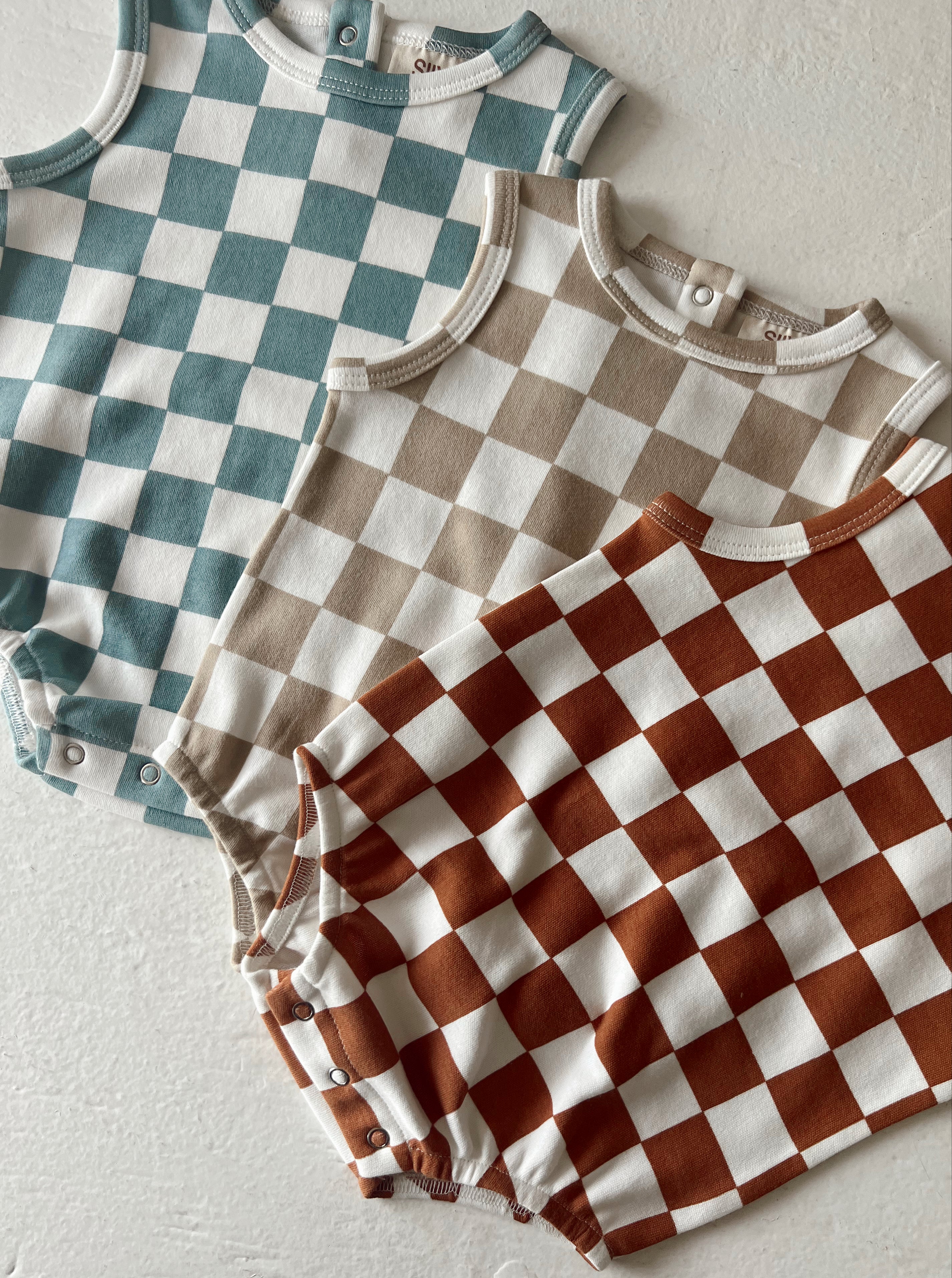 Three checkered baby rompers in teal, beige, and rust colors on a light background.