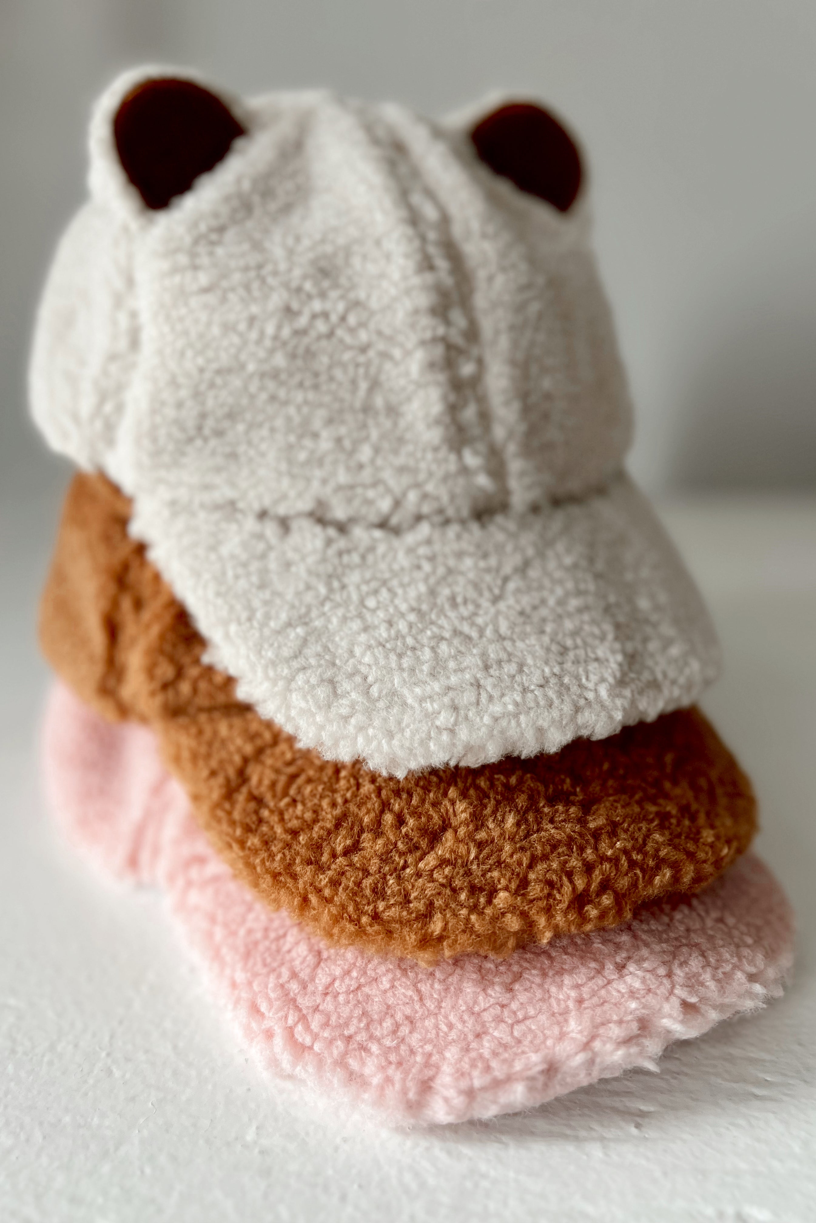 Fuzzy bear-themed cap stacked on soft, colorful fleece hats in shades of brown and pink against a neutral background.