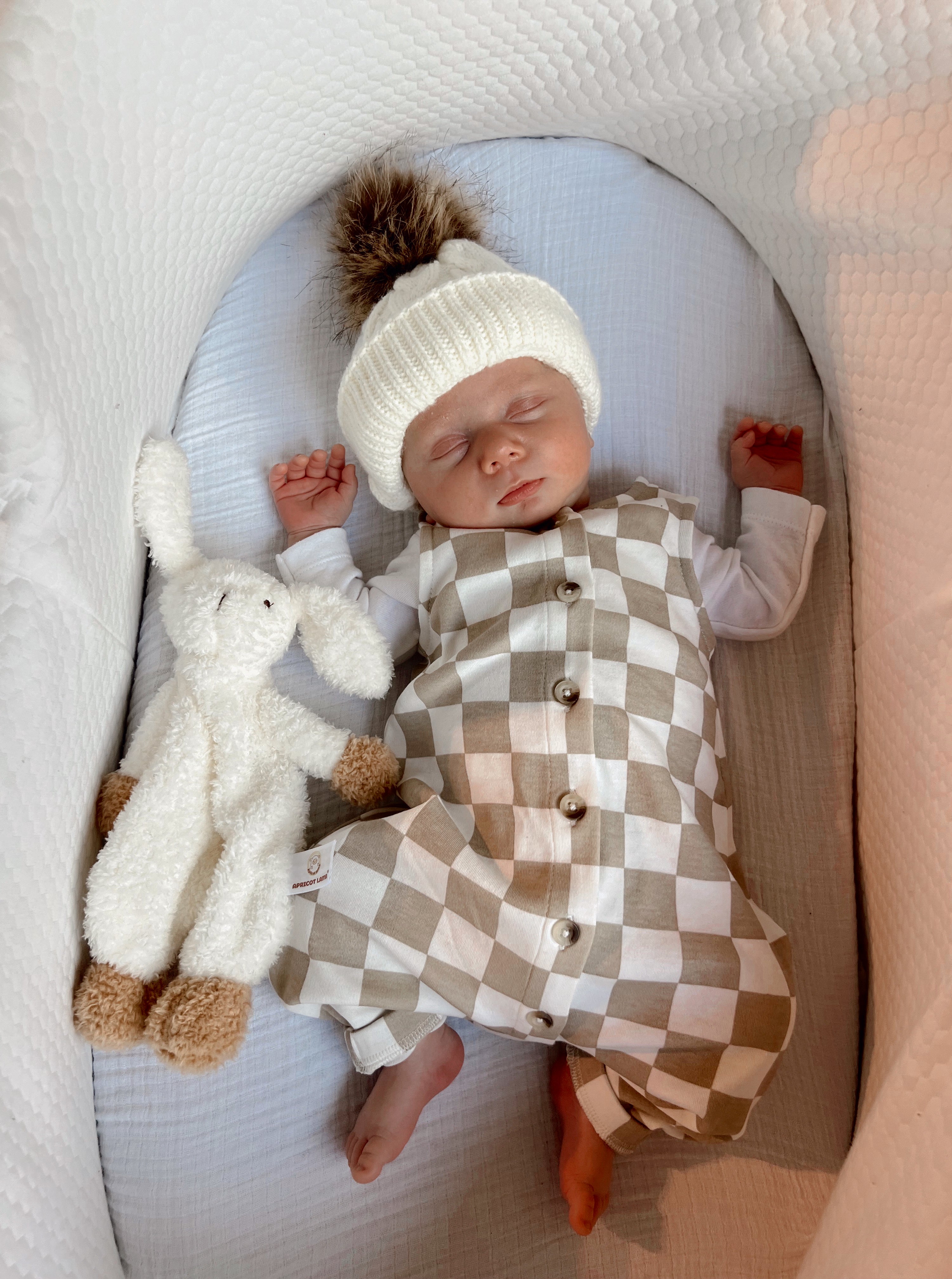 Baby in a checkered outfit and knit hat sleeps peacefully beside a soft toy in a cozy bassinet.