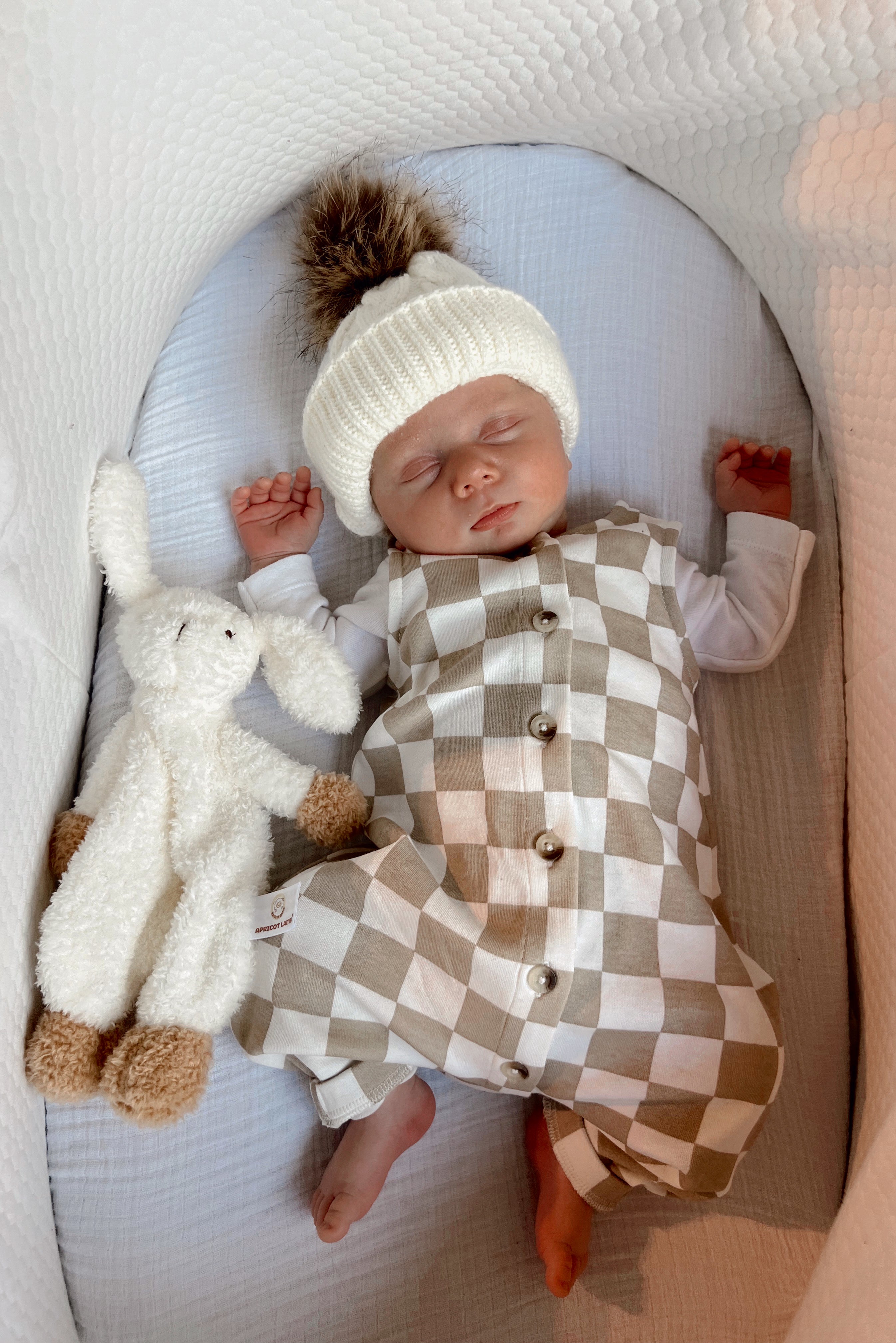 Baby in a checkered outfit and knit hat sleeps peacefully beside a soft toy in a cozy bassinet.
